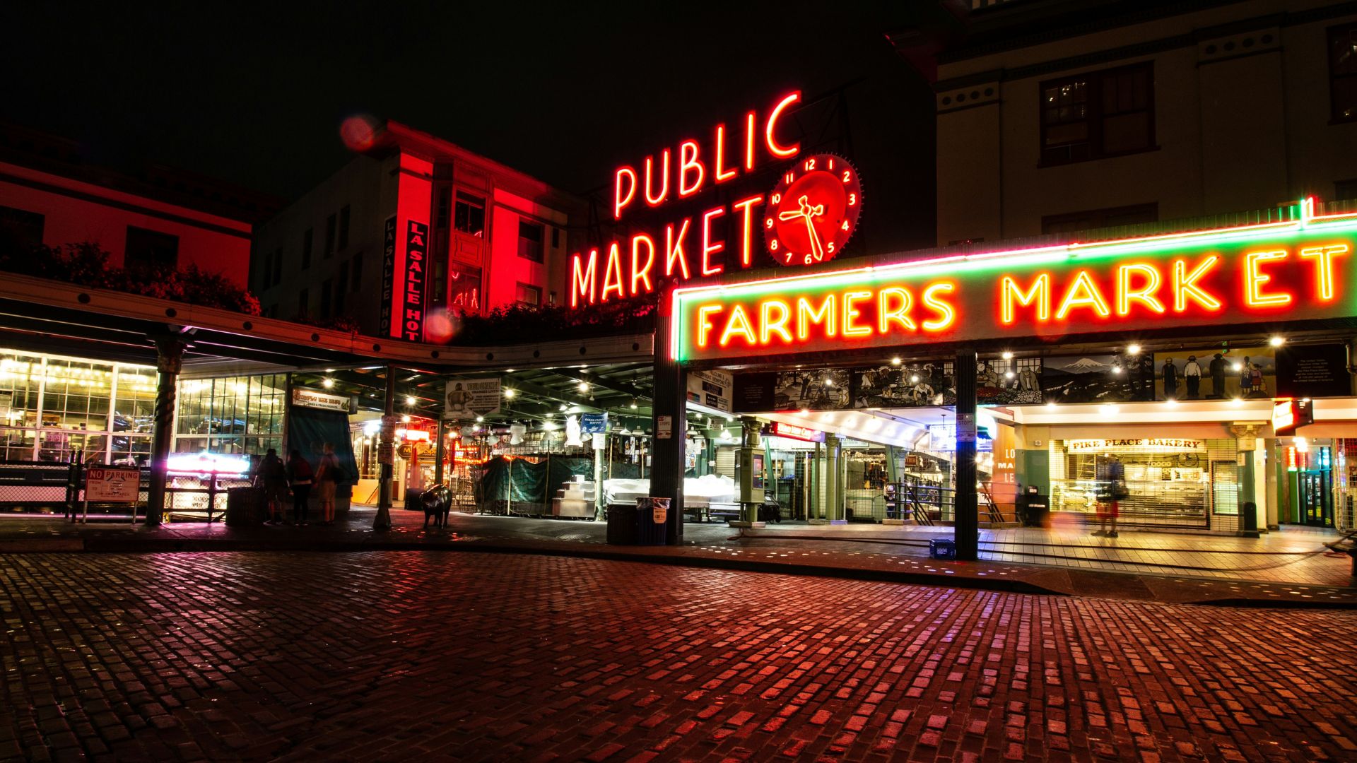 A panoramic nighttime view of the iconic neon "Public Market" and "Farmers Market" signs glowing red and green above a brick-paved street.