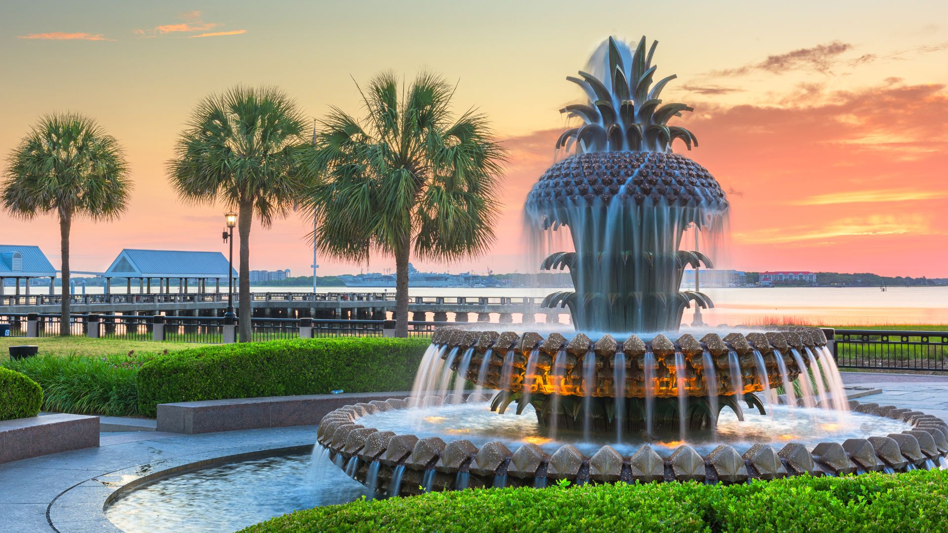 A large, illuminated pineapple-shaped fountain in Waterfront Park with palm trees and a wooden pier in the background, set against a vibrant orange and pink sunset sky over the water.