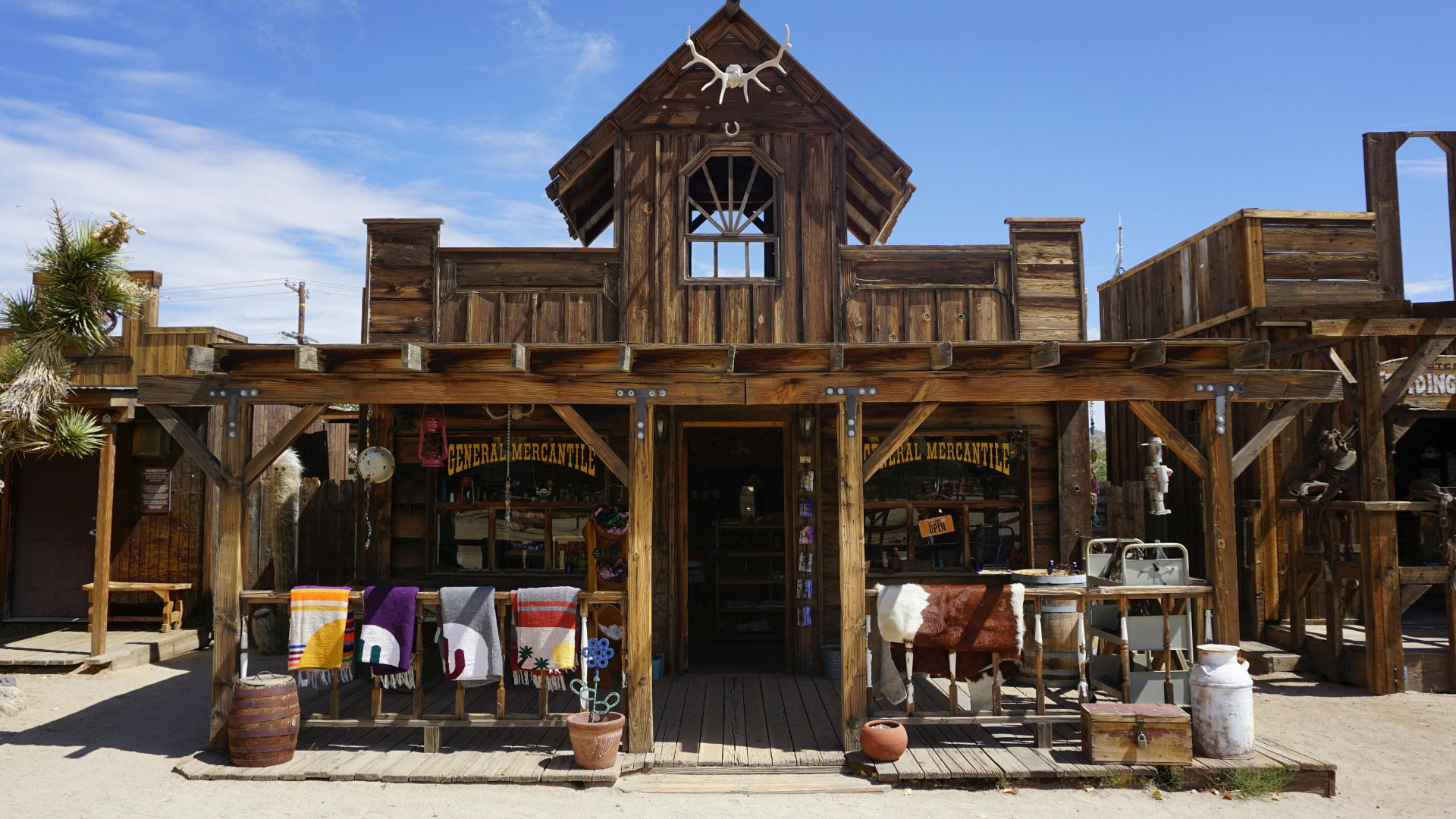 A two-story, rustic wooden general store building in the old Western-style movie set of Pioneertown, California, with a covered porch displaying colorful blankets, an animal skull mounted on the peak of the roof, and sandy ground in the foreground.