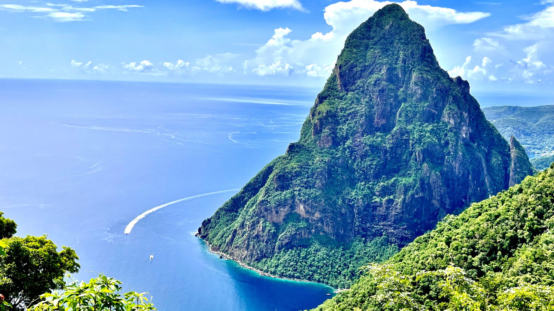 A large, lush green volcanic mountain rises steeply from the blue ocean under a partly cloudy sky, with a boat leaving a white wake in the water.