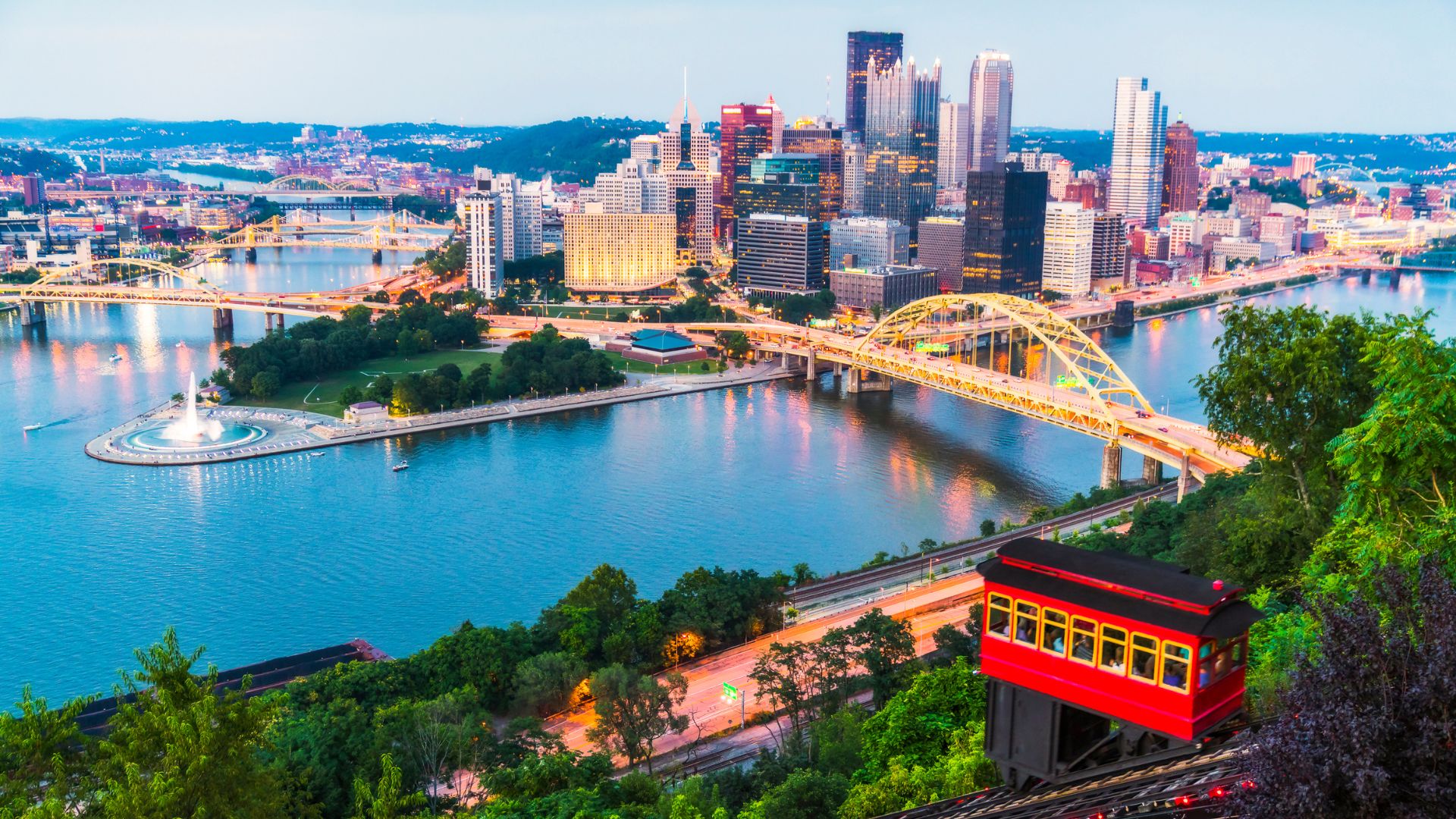 A panoramic, evening view of the Pittsburgh skyline featuring skyscrapers, numerous bridges including a yellow arch bridge, and a river fountain at the confluence of three rivers. A red funicular car is visible on the tracks in the wooded foreground.
