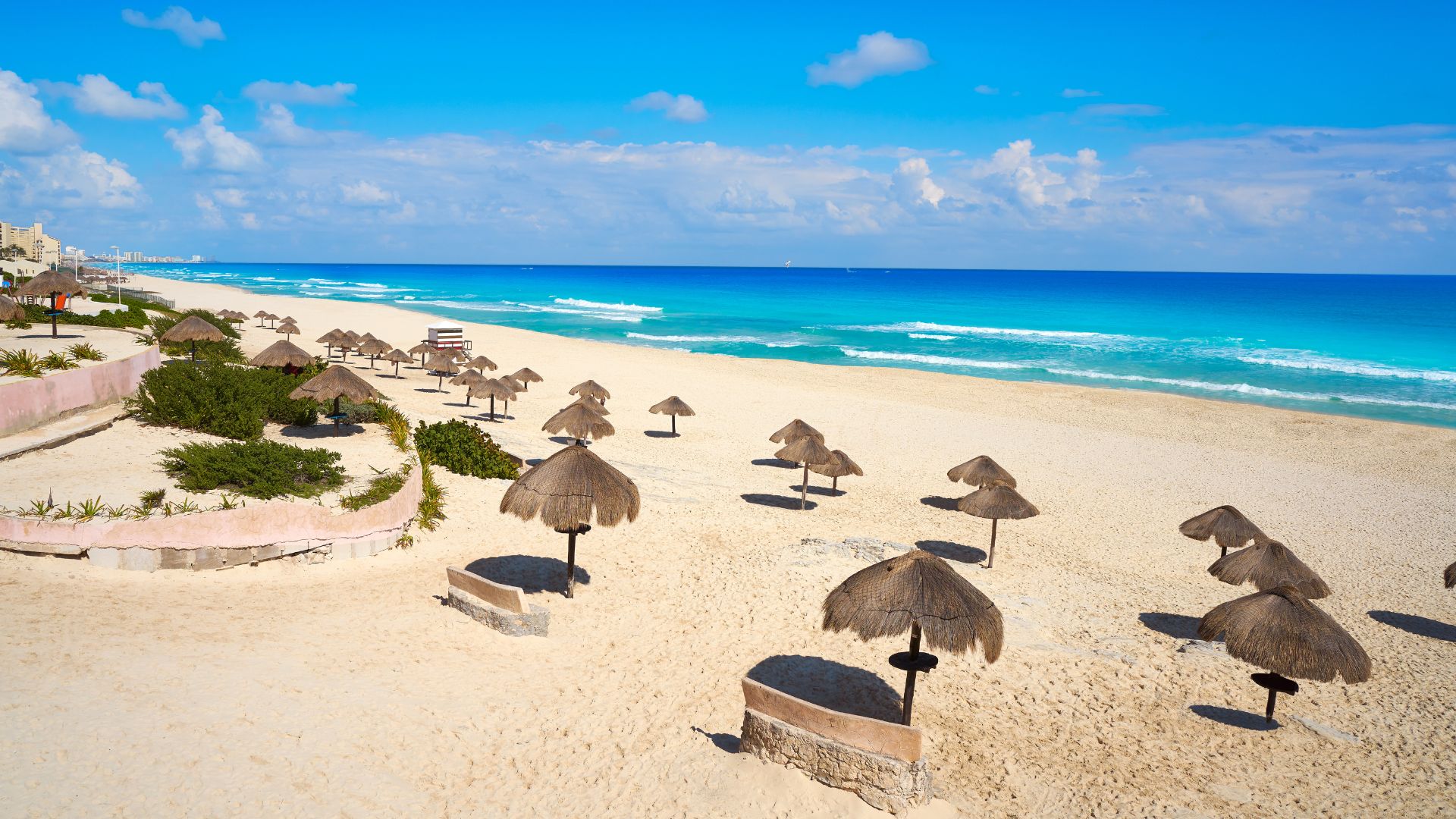 A sunny beach with white sand, turquoise water, and several thatched umbrellas (palapas).