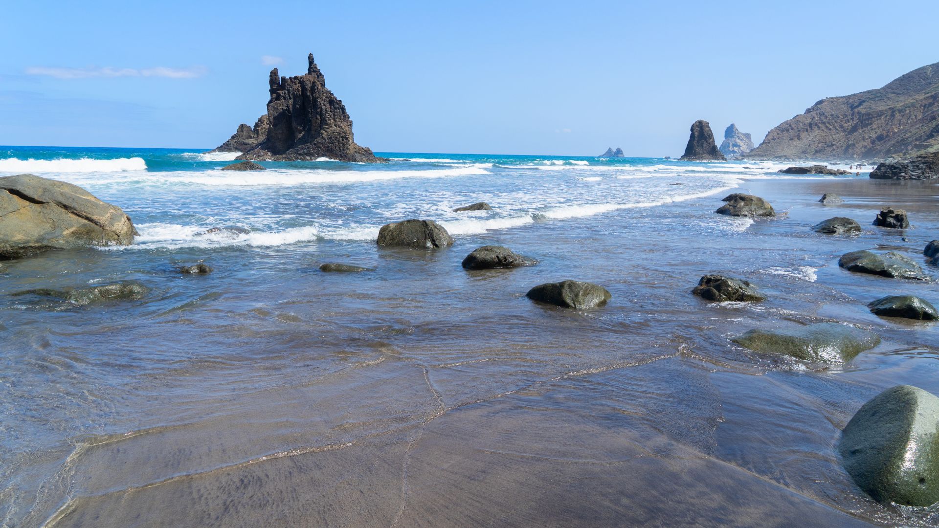 A scenic view of Playa de Benijo beach in Tenerife, featuring black sand, large volcanic rock formations in the turquoise ocean, and waves washing ashore under a clear blue sky.