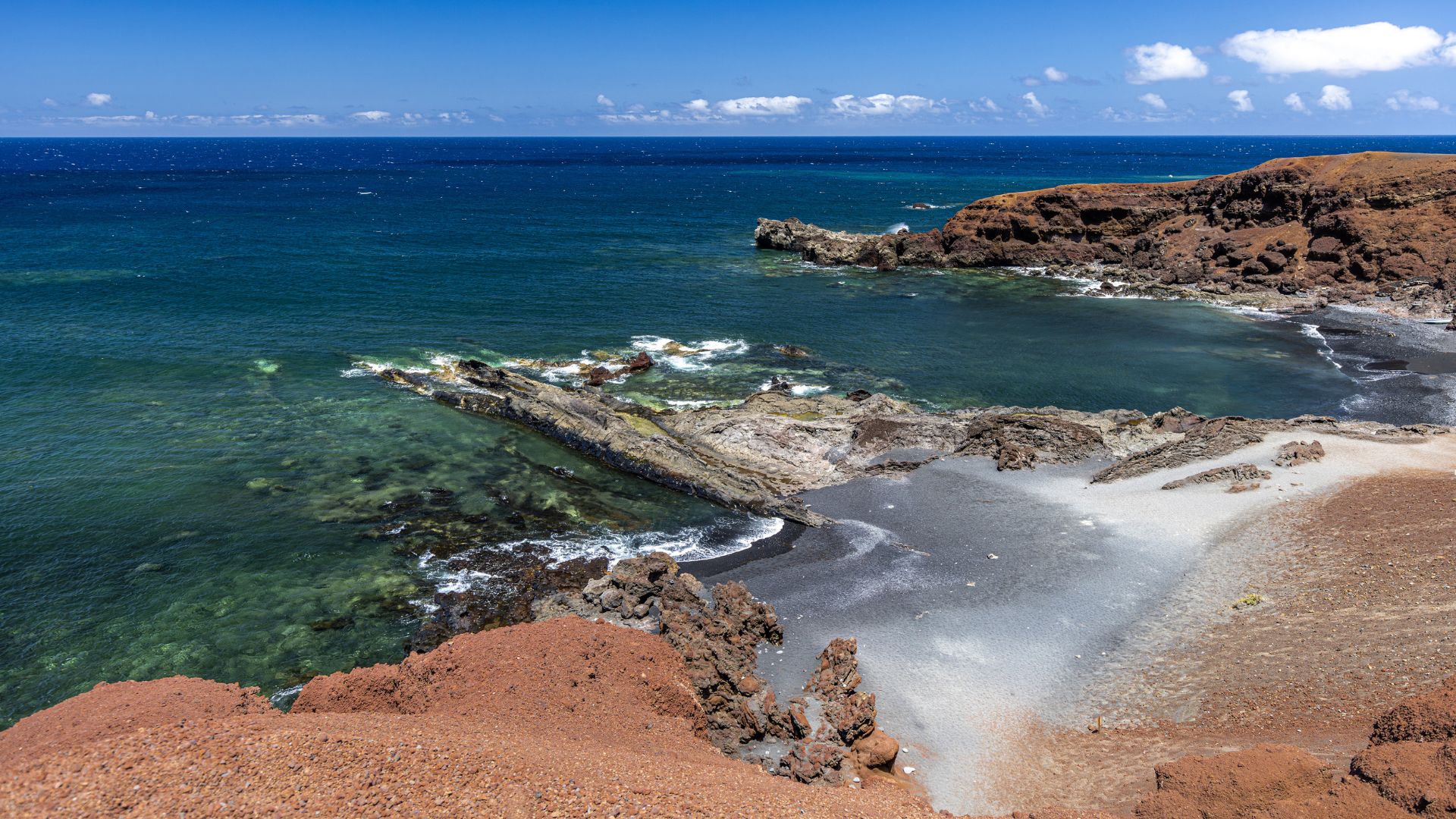 A dramatic coastal view of El Golfo, Lanzarote, featuring dark sand, red volcanic cliffs, and the blue Atlantic Ocean