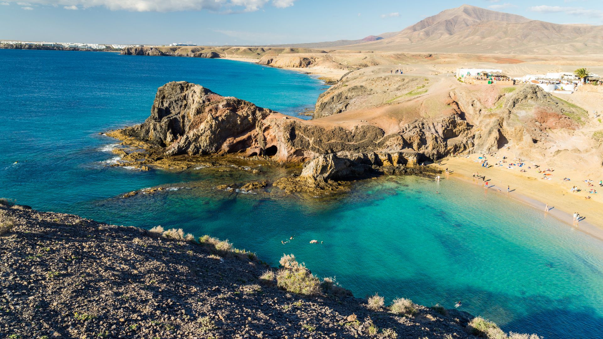 A high-angle photo of a secluded golden sand beach with turquoise water, surrounded by arid, rocky cliffs and a large rock formation in the water.