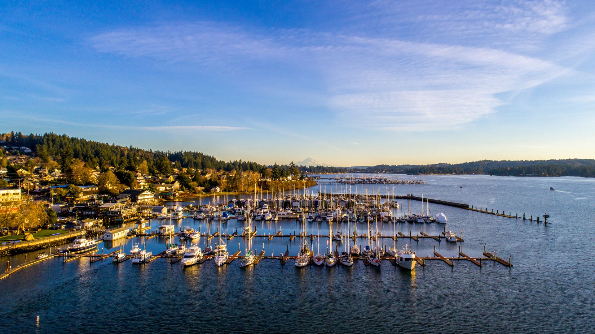 An aerial, panoramic daytime view of the Poulsbo, Washington marina filled with sailboats and docks, the water of Liberty Bay, and residential houses dotting the lush green hillside under a blue sky with light clouds.