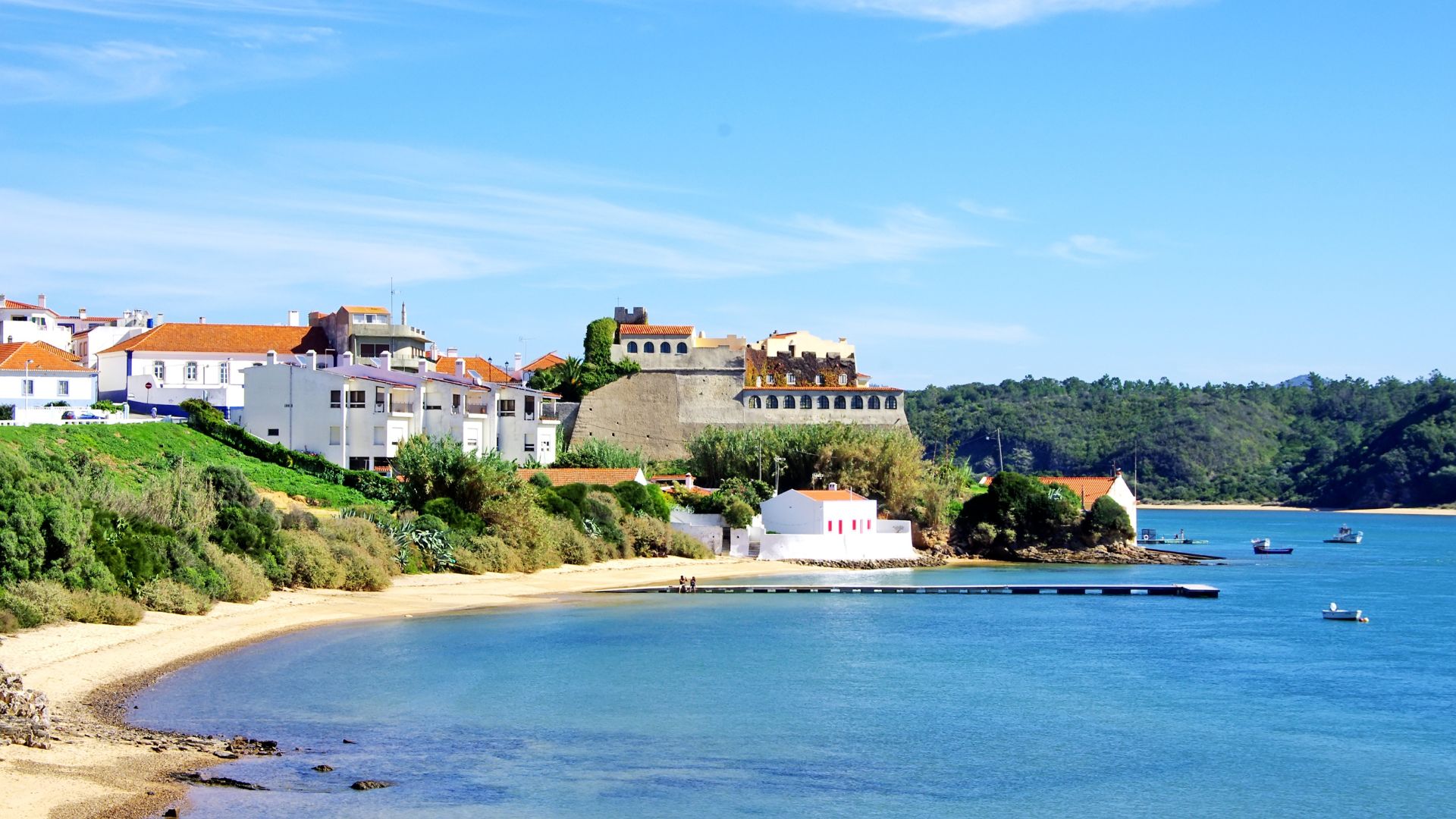 A sunny view of the sheltered Praia da Franquia beach in Vila Nova de Milfontes, Portugal, featuring the historic Forte de São Clemente on an embankment by the Mira River estuary and buildings of the town on the hillside.
