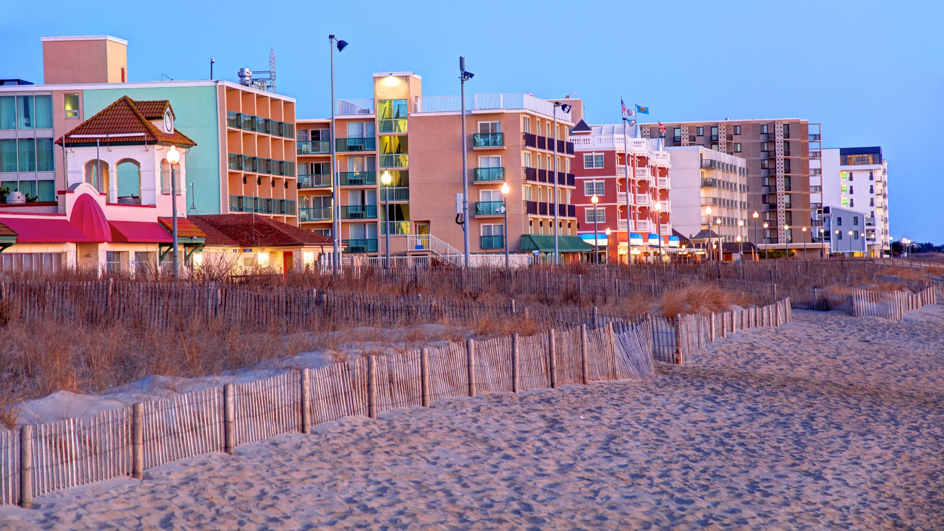 A sandy beach with low grass dunes and long wooden fences leading towards a dense row of multi-story beachfront hotels and resort buildings at dusk.