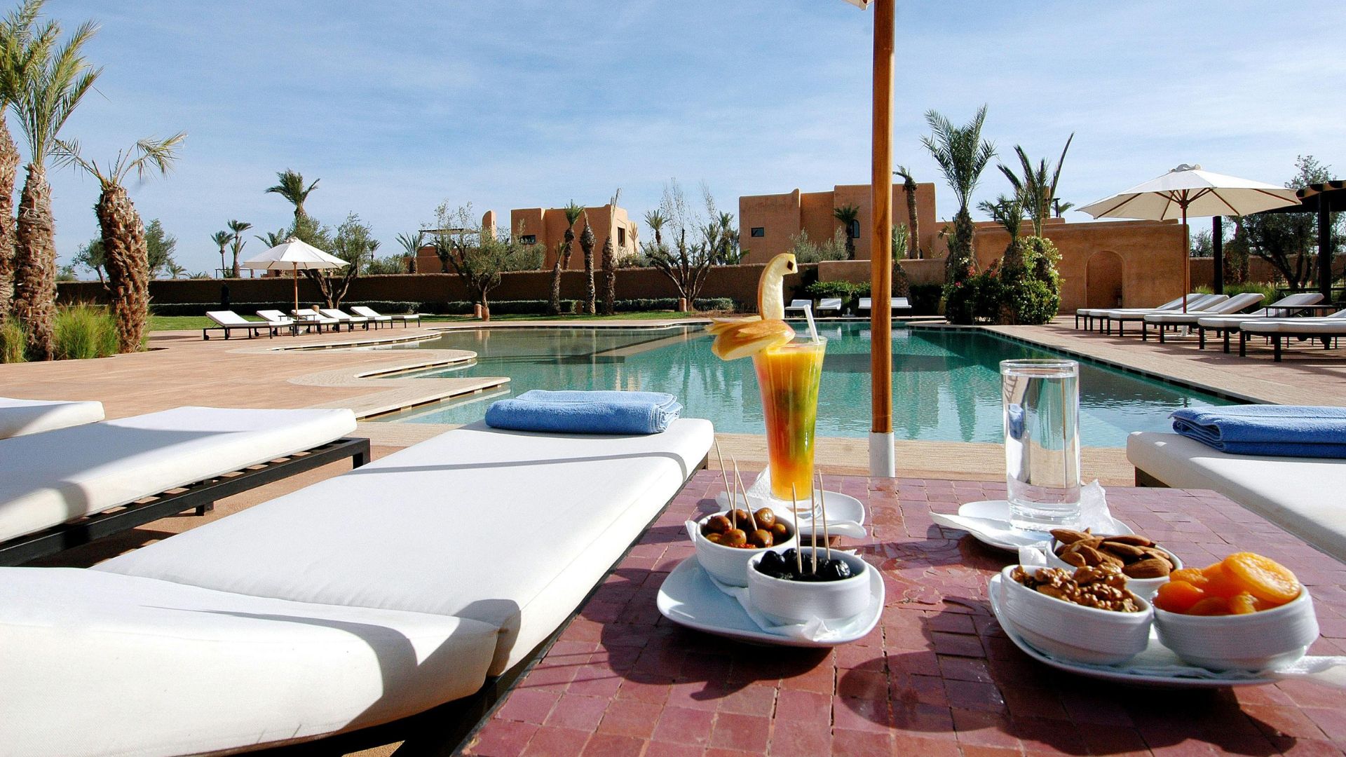A sunny outdoor resort pool area with white lounge chairs, snacks, drinks, and traditional-style buildings in the background.