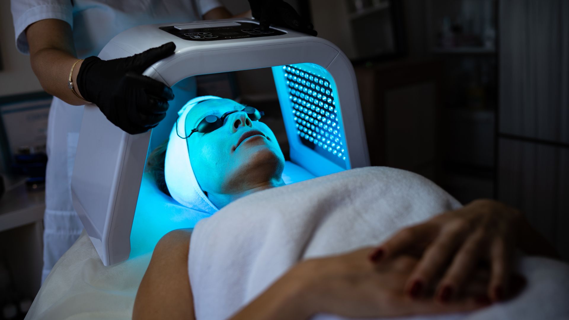 A person lies in a spa setting undergoing a blue LED light therapy facial treatment, wearing protective eye covers. A technician wearing black gloves adjusts the overhead light panel.