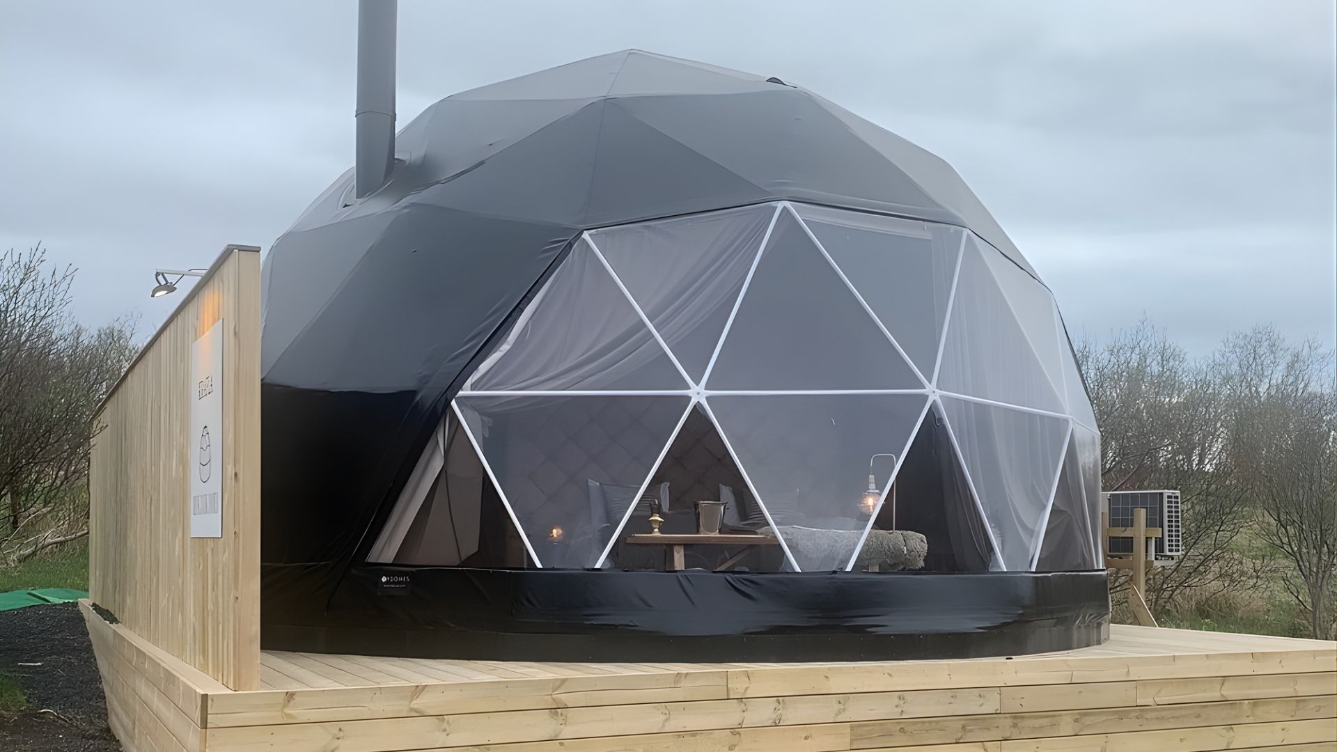 A large, black, dome-shaped glamping tent with white geodesic framework is set on a wooden deck in a grassy field on an overcast day.
