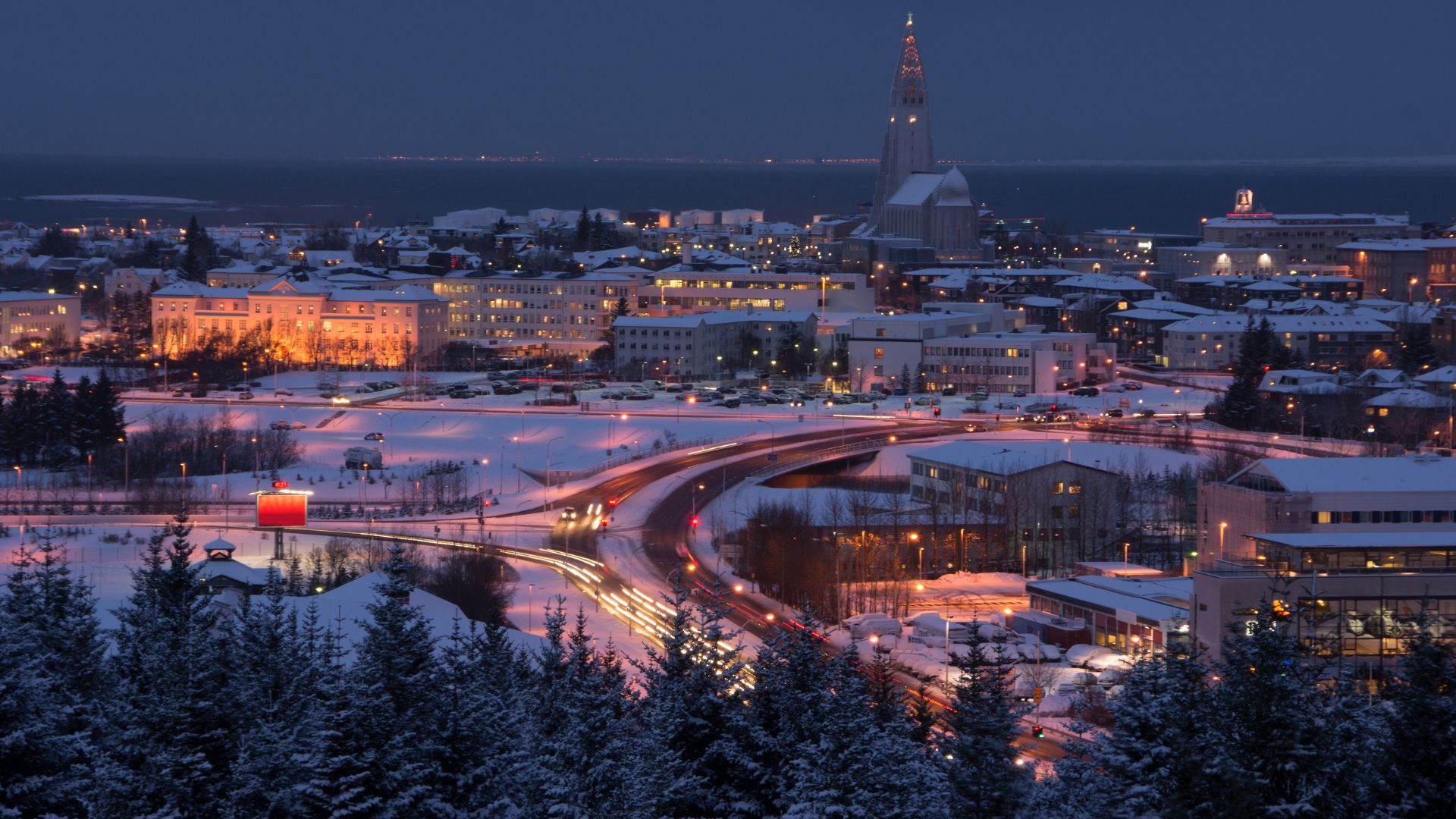 A night-time aerial photograph of the snow-covered city of Reykjavík, Iceland, with the illuminated Hallgrímskirkja church tower standing tall above the city lights and snowy evergreen trees in the foreground.