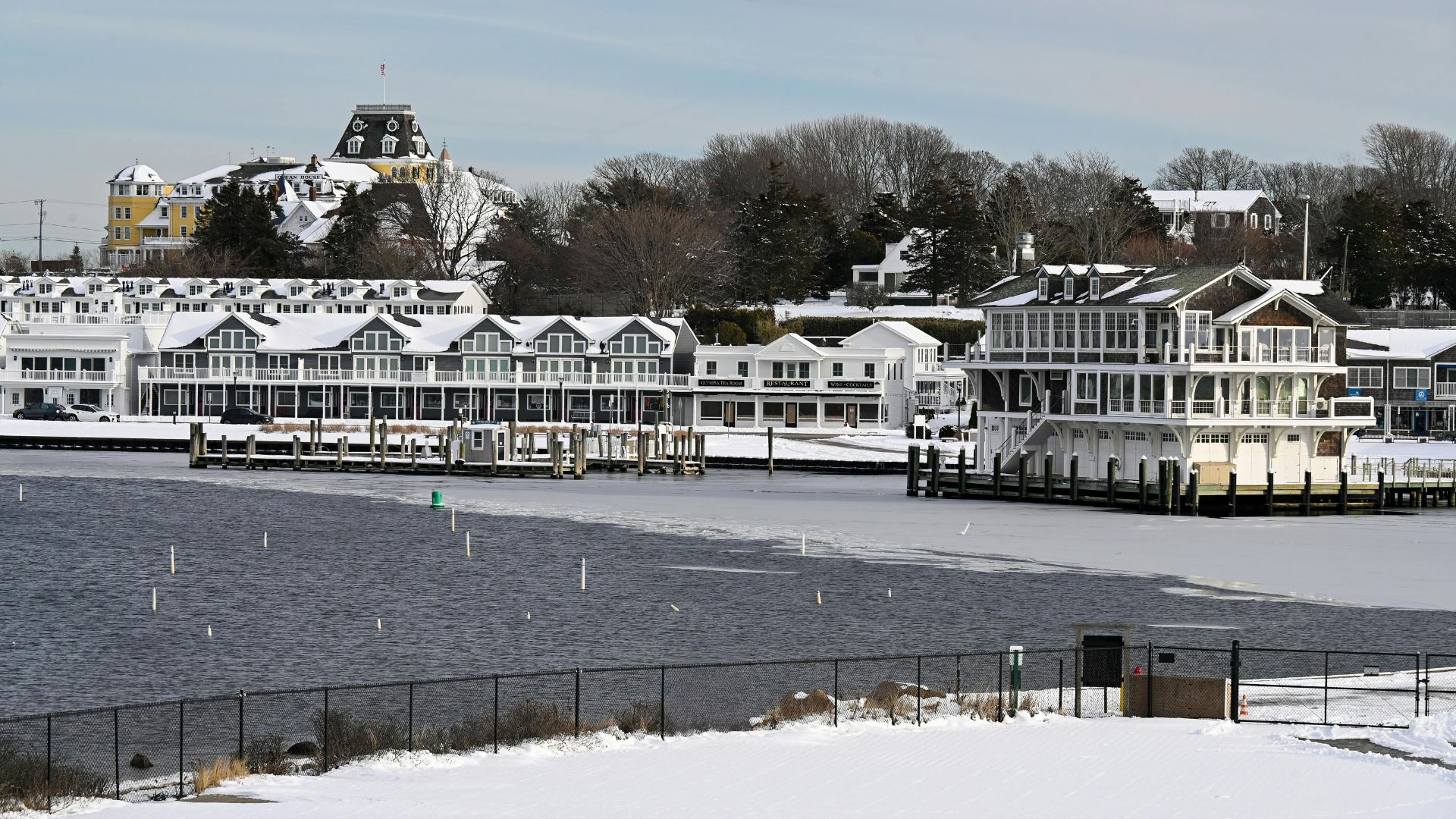A snowy coastal scene in New England features several large white waterfront buildings and a prominent yellow historic hotel perched on a hill overlooking a partially frozen harbor.