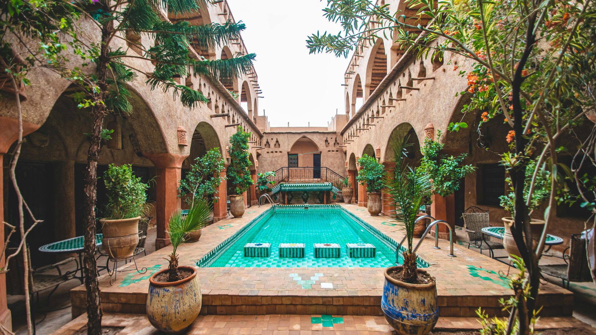 A tranquil internal courtyard of a traditional Moroccan riad featuring a long, narrow swimming pool, lush potted plants, arched walkways, and tiled flooring.
