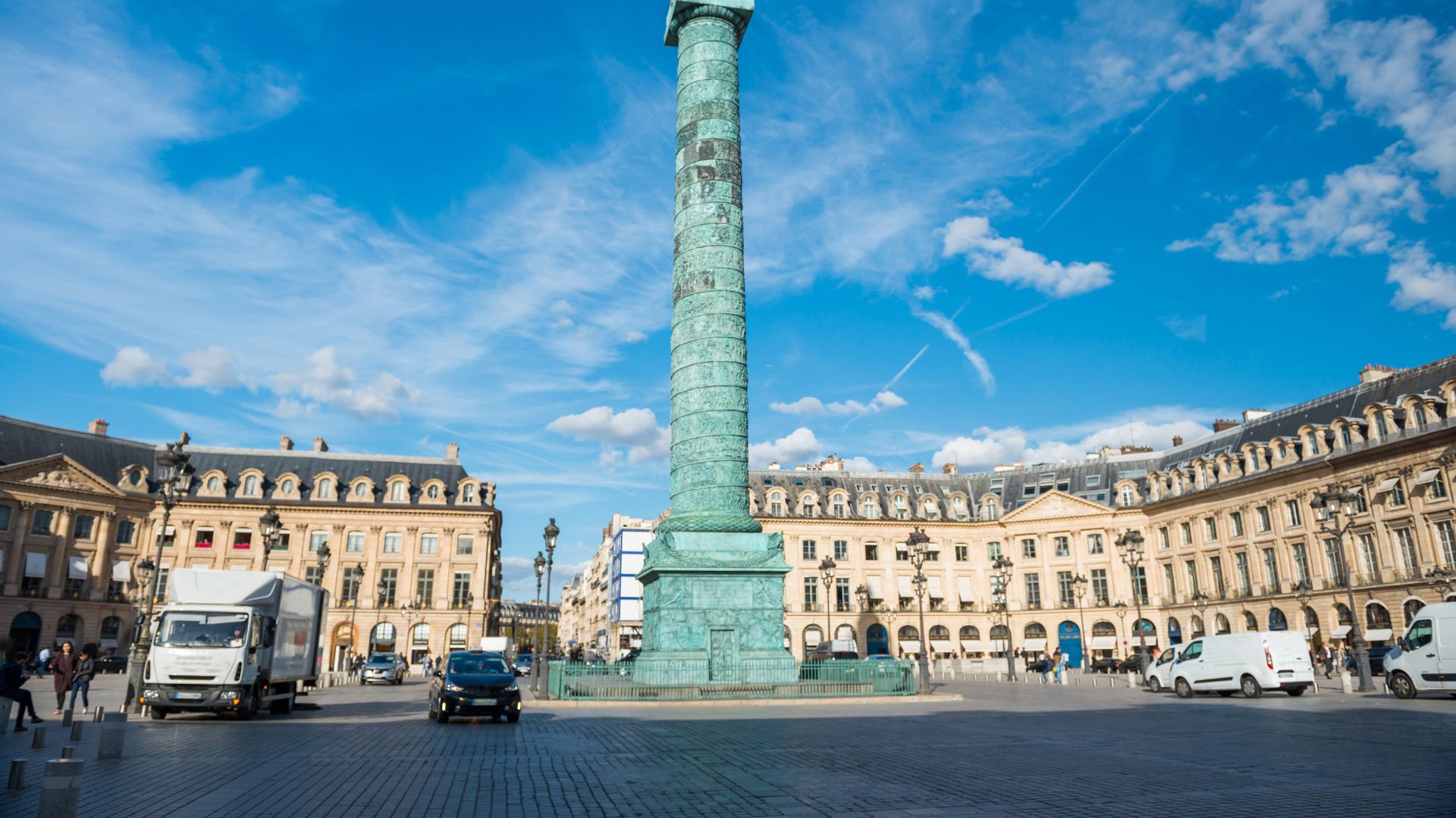 A panoramic view of the Place Vendôme in Paris, featuring the central green Vendôme Column, surrounding classical buildings, and vehicles on a sunny day.