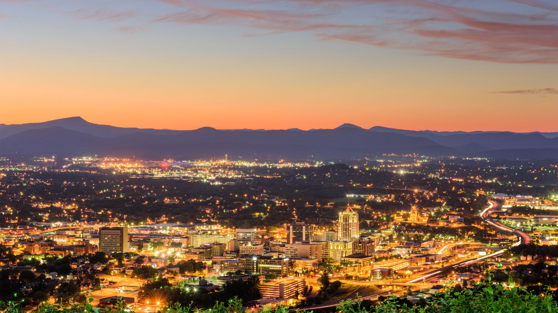 A panoramic, elevated view of the Roanoke, Virginia, city lights illuminated at dusk, set against a backdrop of dark Blue Ridge Mountains.