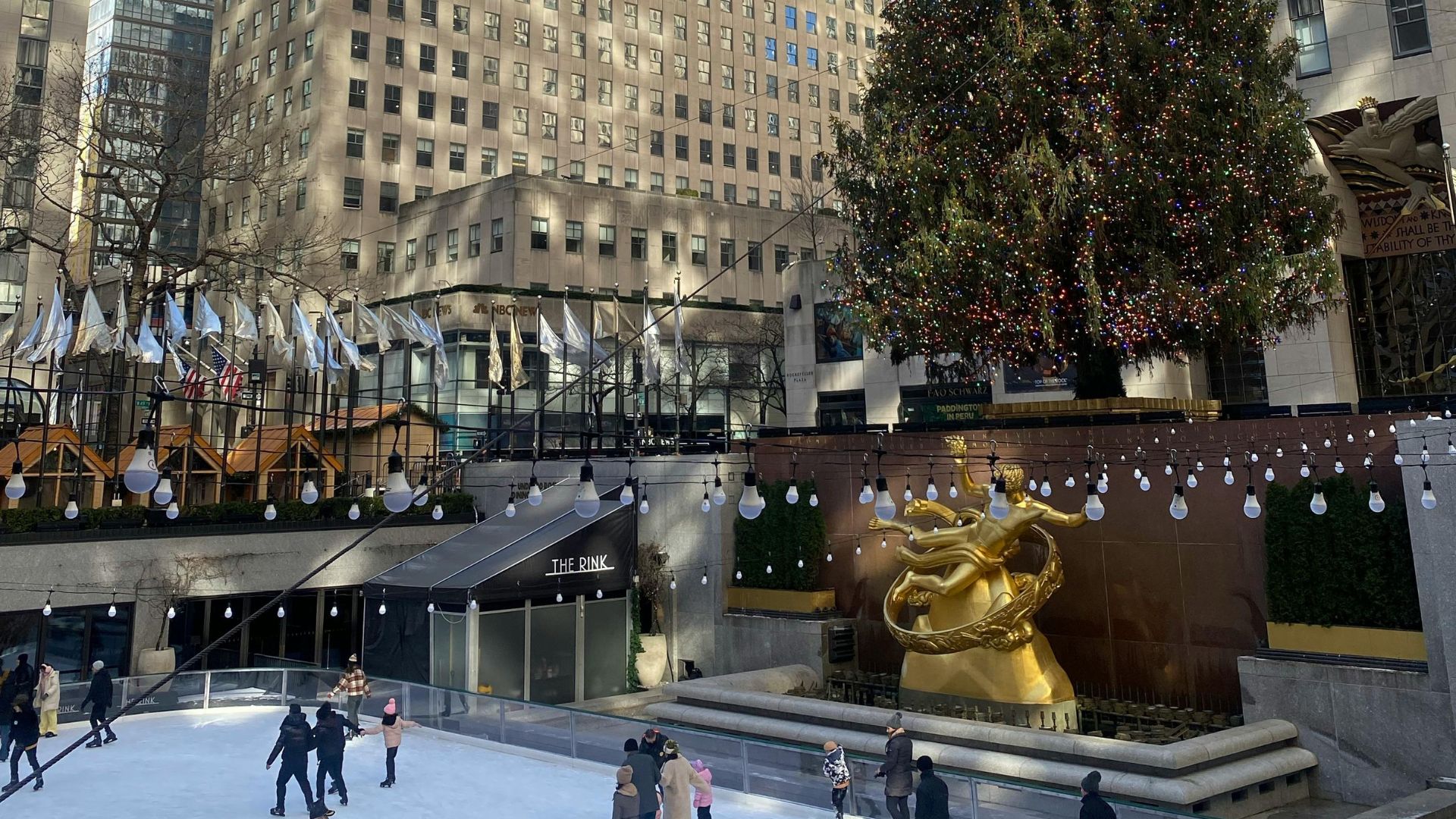 A view of the outdoor ice skating rink and golden Prometheus statue at Rockefeller Center in Midtown Manhattan, New York City, surrounded by buildings and international flags.