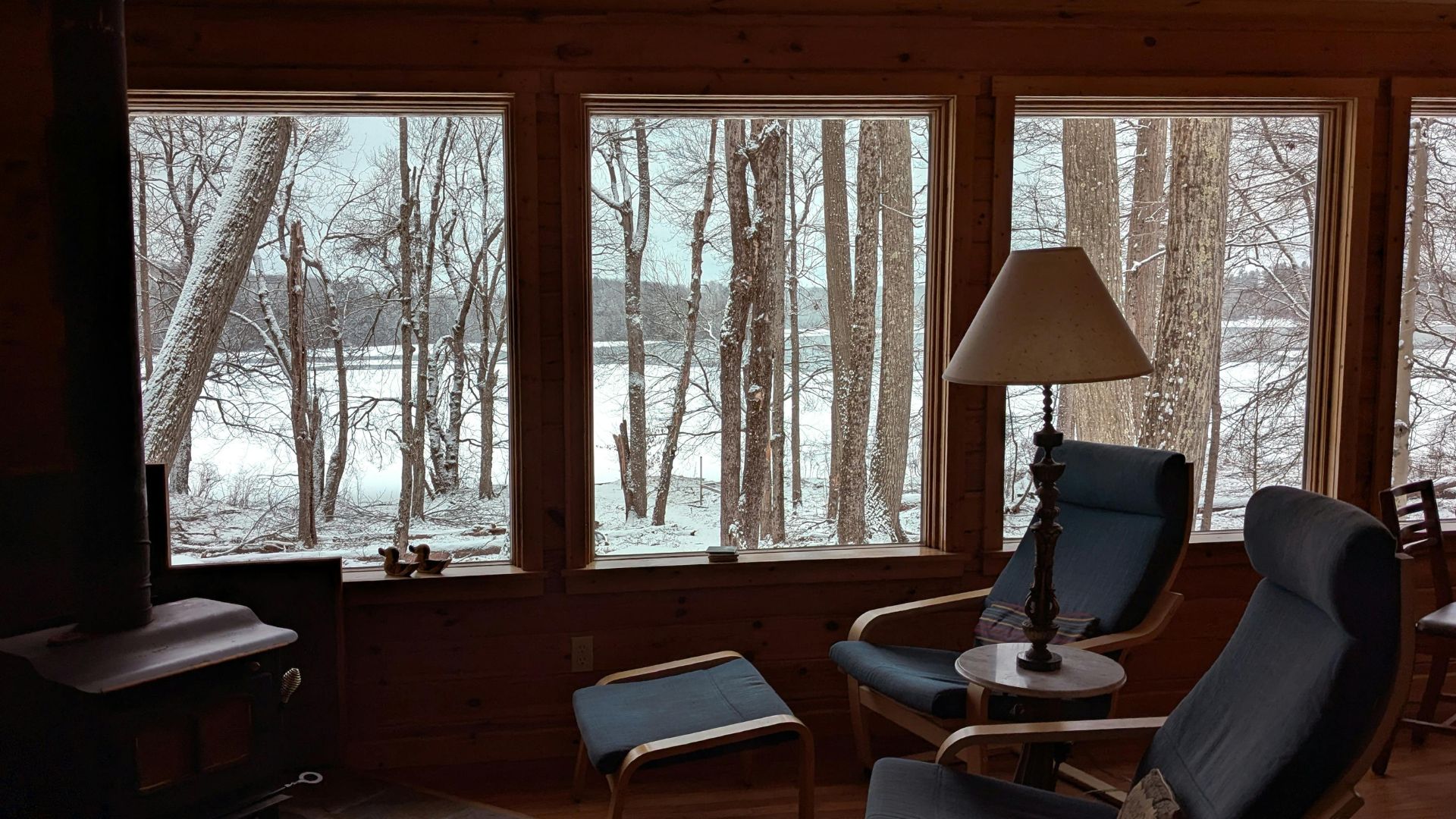 A cozy cabin living room with wood-paneled walls and large windows overlooks a snowy, forested landscape and frozen lake, featuring blue accent chairs and a floor lamp.