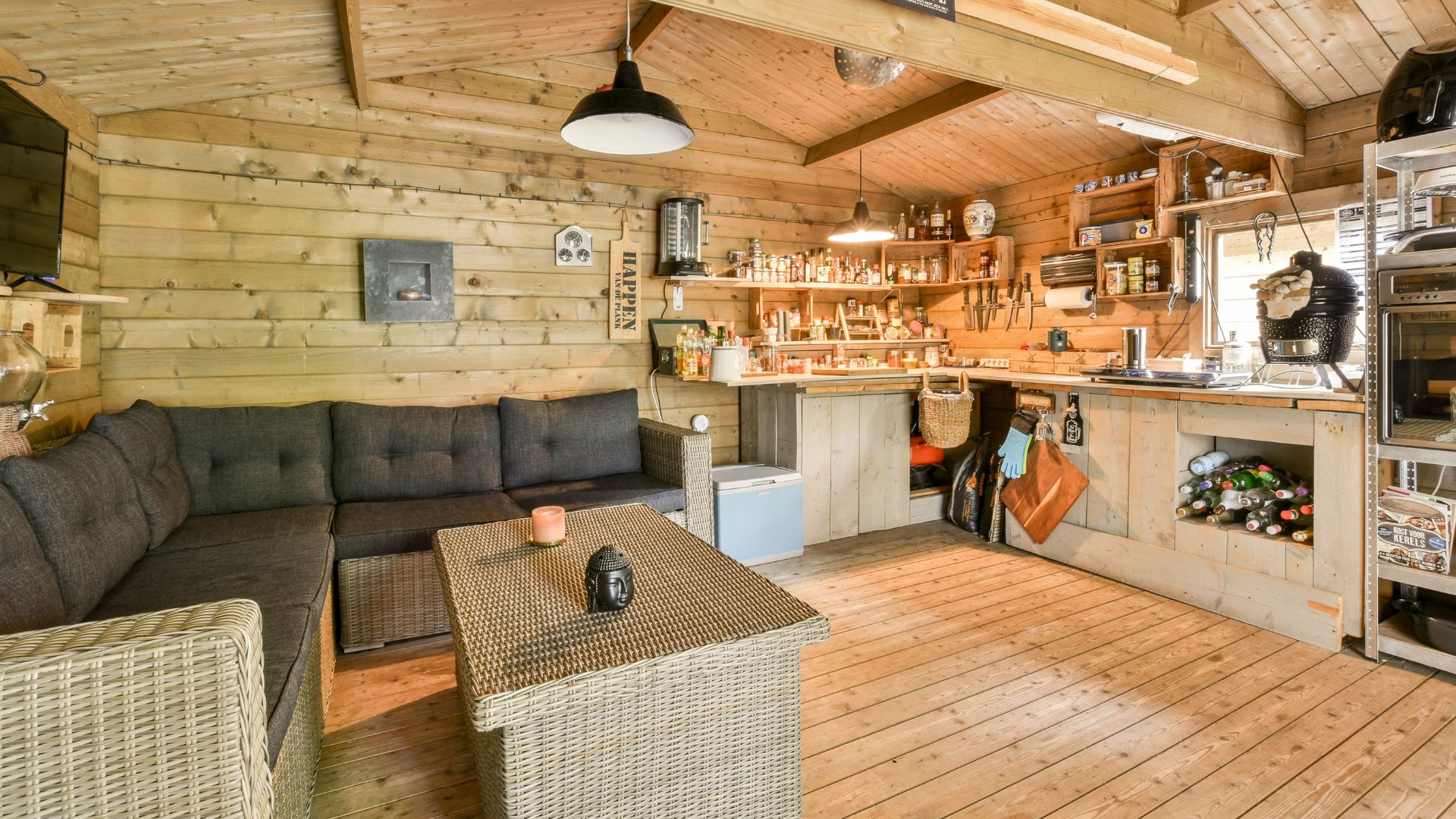 The interior of a rustic wooden cabin featuring an L-shaped gray sofa, a woven coffee table, and an open kitchenette area with a black pendant lamp hanging over the counter.