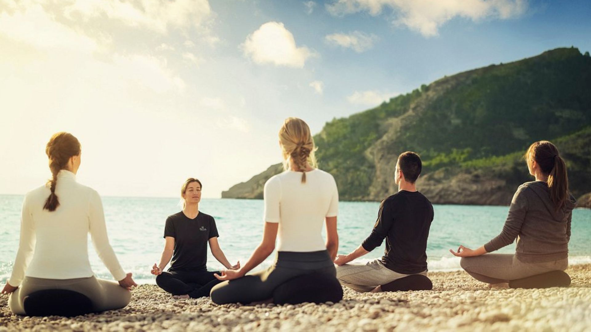 The image depicts a group of people doing yoga or meditating on a pebble beach, with the ocean in front of them and a large, green, tree-covered hill or mountain in the background.