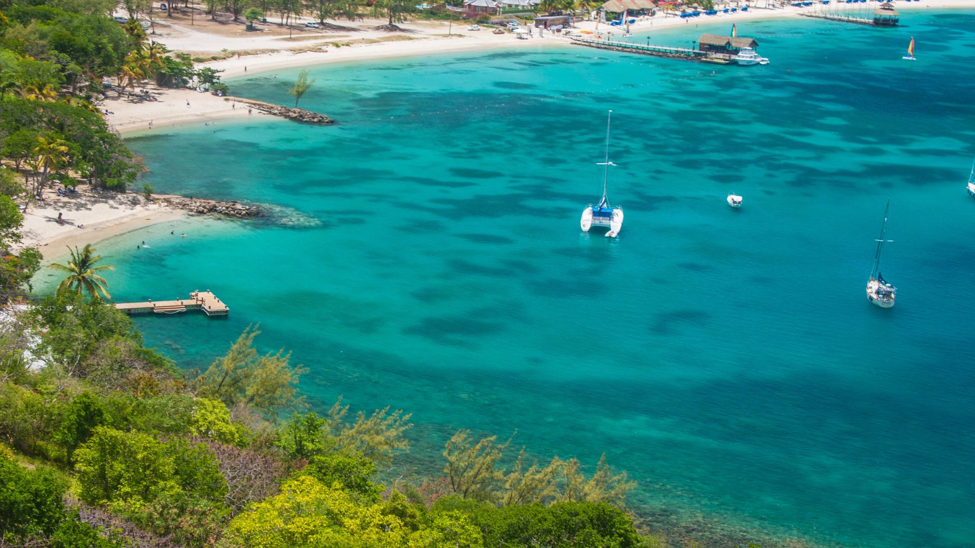 An aerial view of a tropical bay in St. Lucia featuring turquoise water, a sandy beach, boats, and lush green hillsides.
