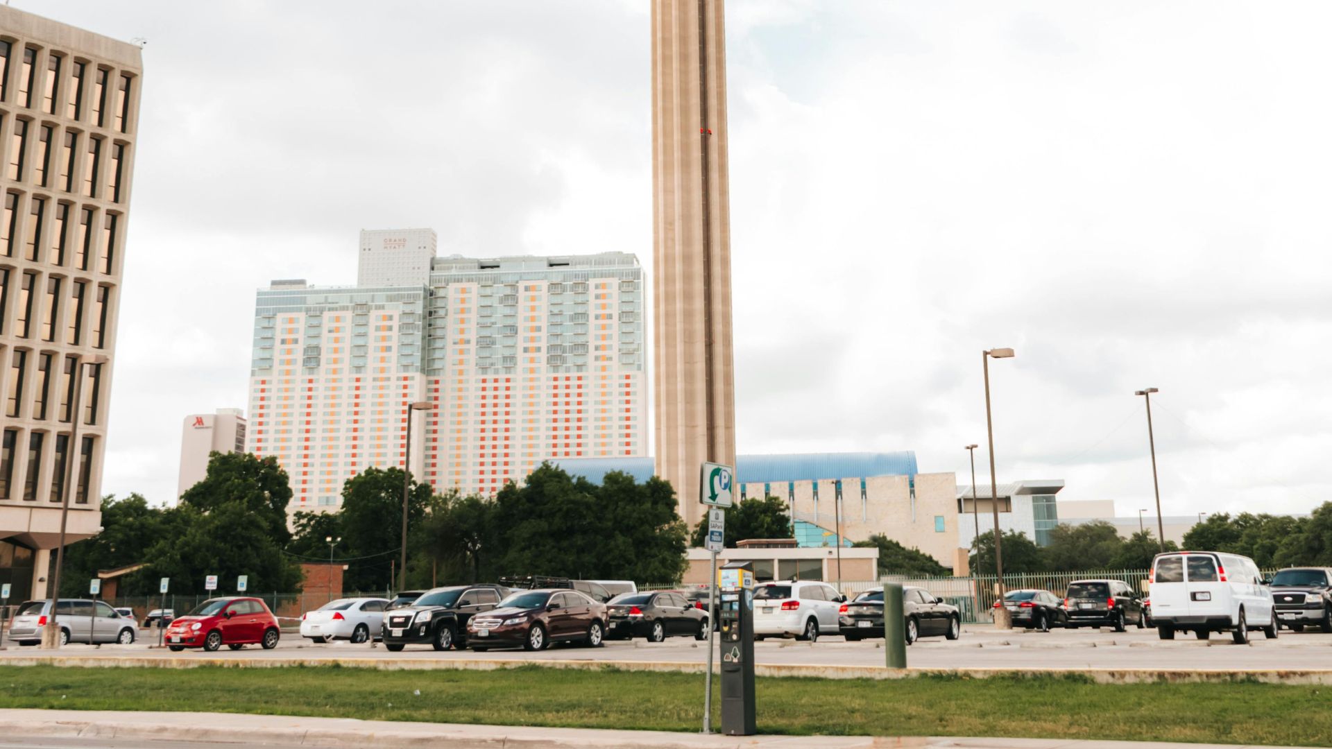 A wide view of the Tower of the Americas in San Antonio, Texas, featuring its tall central column and circular top against a blue sky with white clouds, with surrounding buildings and a street of cars in the foreground.