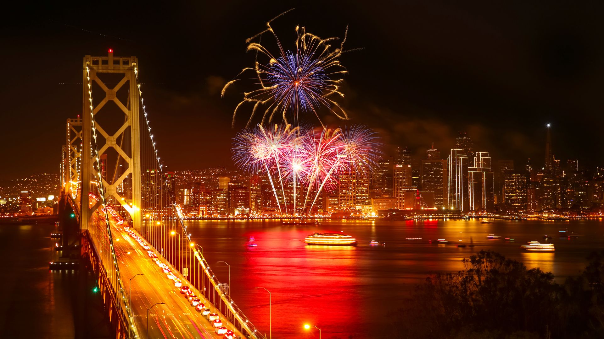 A view across the San Francisco Bay at night with a red light reflection on the water, featuring a traffic-filled Bay Bridge on the left, city skyscrapers on the right, and a vibrant fireworks display over the water.