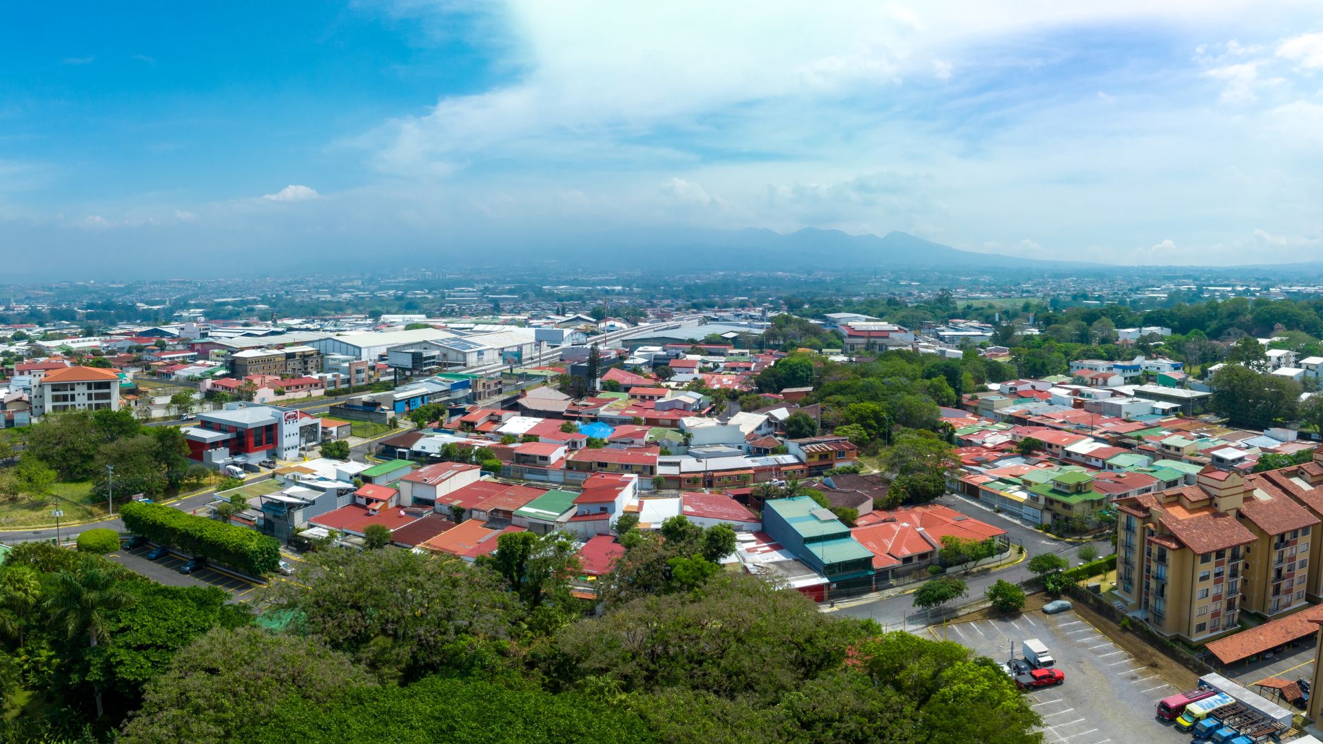 An aerial photo showcasing a dense urban area in San José, Costa Rica, with a mix of residential and commercial buildings under a partly cloudy sky, and mountains in the distance.