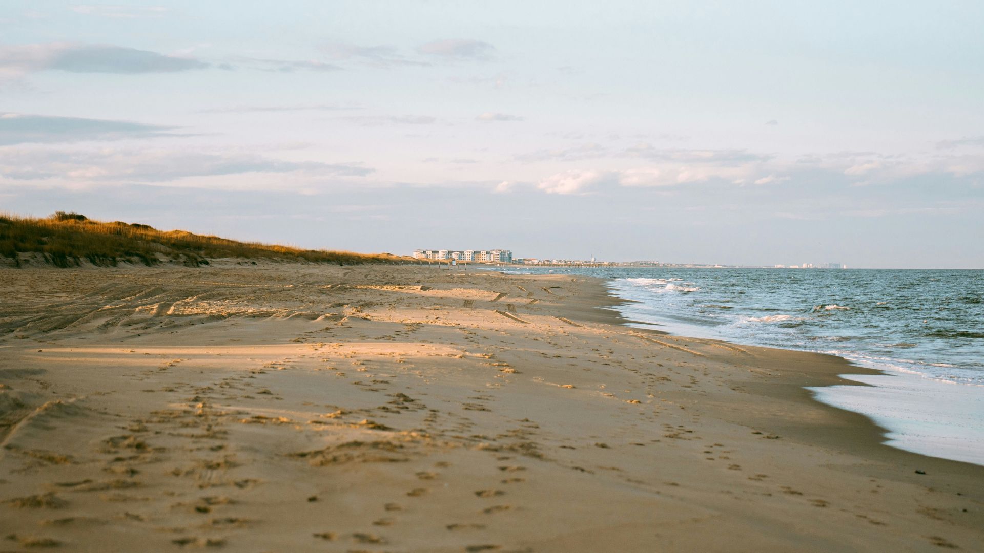 A wide, sandy beach with large dunes covered in tall grass leads to the ocean, with the faint outline of a developed city skyline visible on the distant horizon under a pale, partly cloudy sky.
