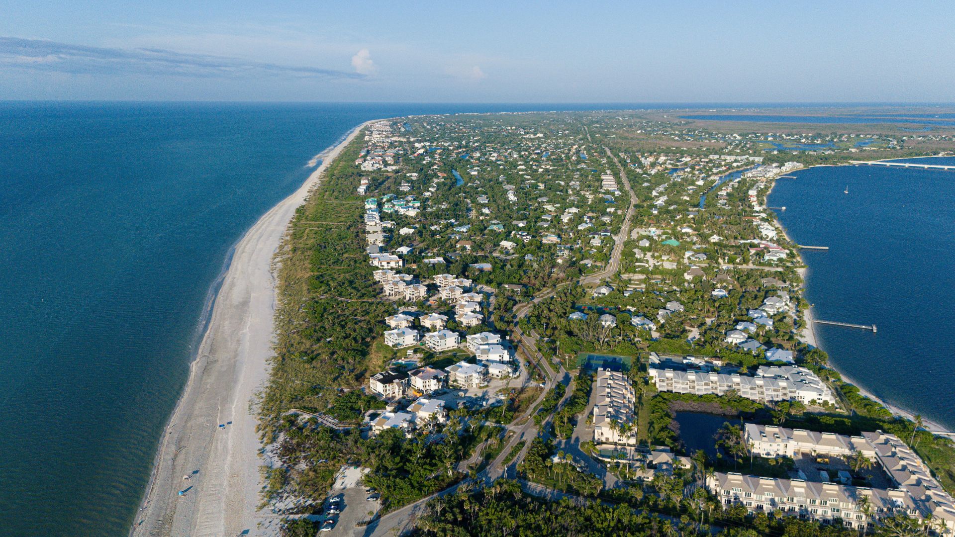 An aerial photo of Sanibel Island, Florida, showcasing a long, narrow barrier island dense with residential homes and tropical vegetation, nestled between the calm, turquoise waters of the Gulf of Mexico and a inland bay.