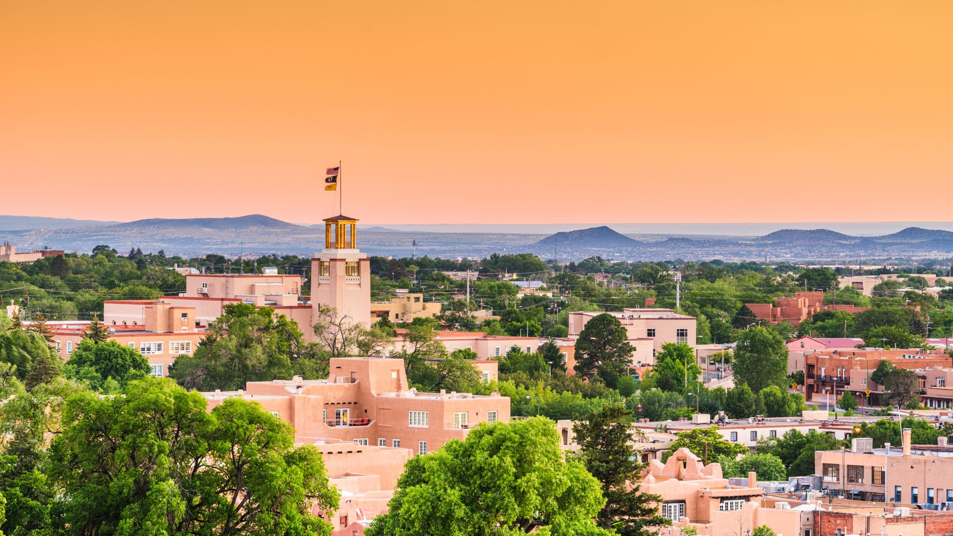 A panoramic view of the city of Santa Fe, New Mexico, featuring its distinctive adobe-style buildings, a central tower with a flag, and mountains in the background under an orange sky.