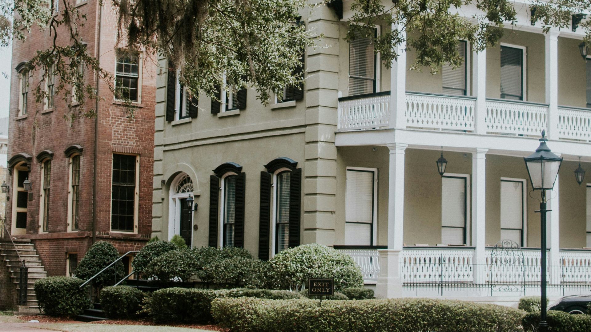 A quiet, brick-paved street in the historic district of Savannah, Georgia, lined with traditional Southern homes featuring multi-story porches and large live oak trees draped in Spanish moss.