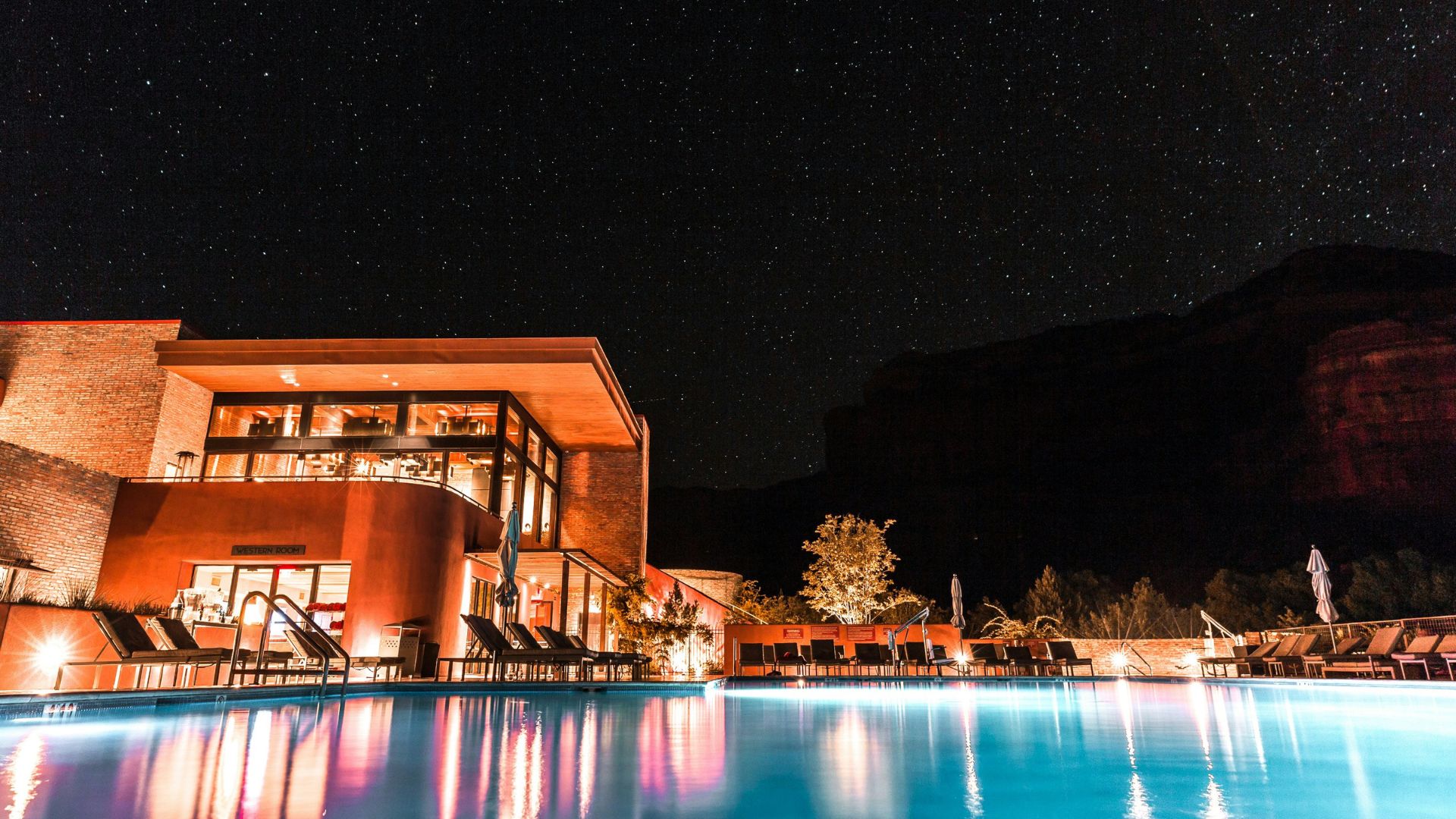 A night time view of a resort's swimming pool reflecting the illuminated buildings, with lounge chairs lining the deck and large, dark red rock mountains visible against a star-filled sky.