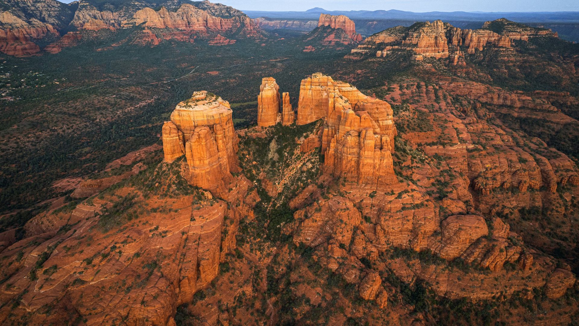 A wide aerial view of the massive red sandstone spires of Cathedral Rock and the surrounding green, rugged desert landscape of Sedona, Arizona, at sunset.