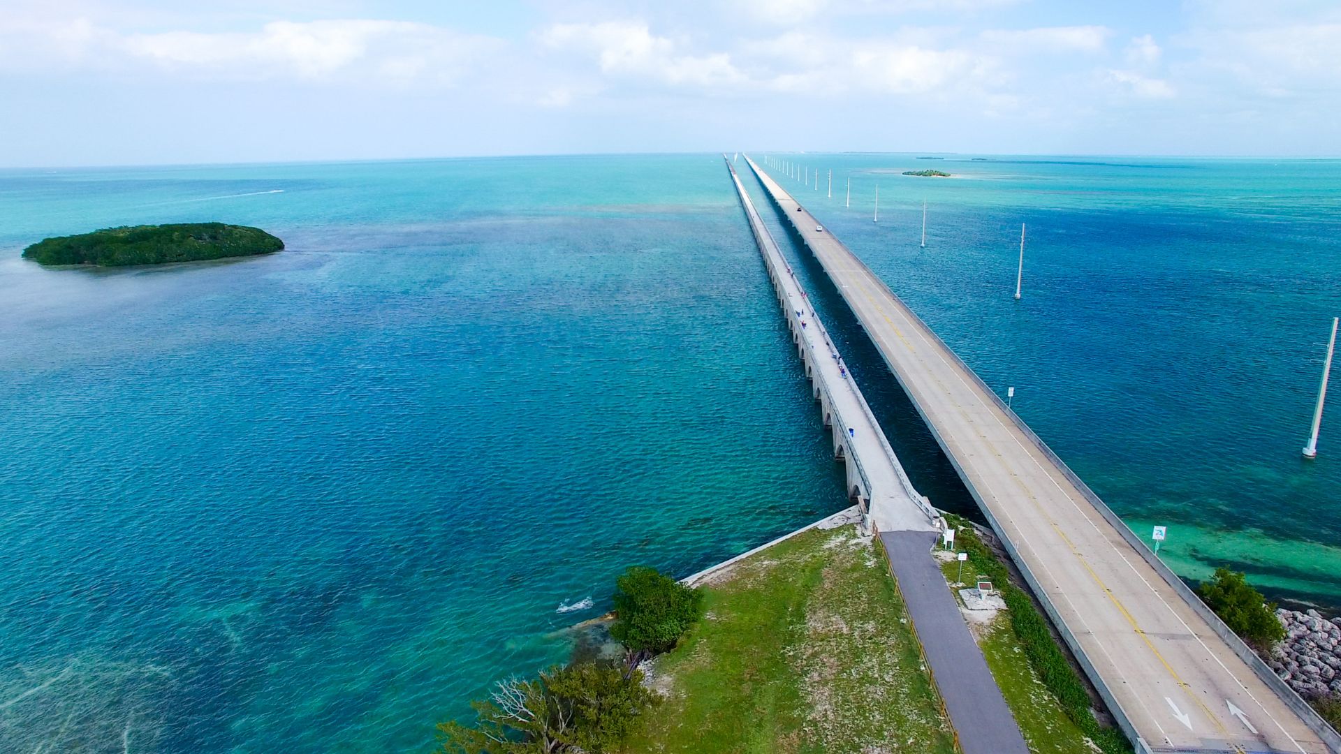 An aerial view of the long Seven Mile Bridge and the parallel old bridge spanning across vibrant turquoise ocean water, with a small green island on the left.