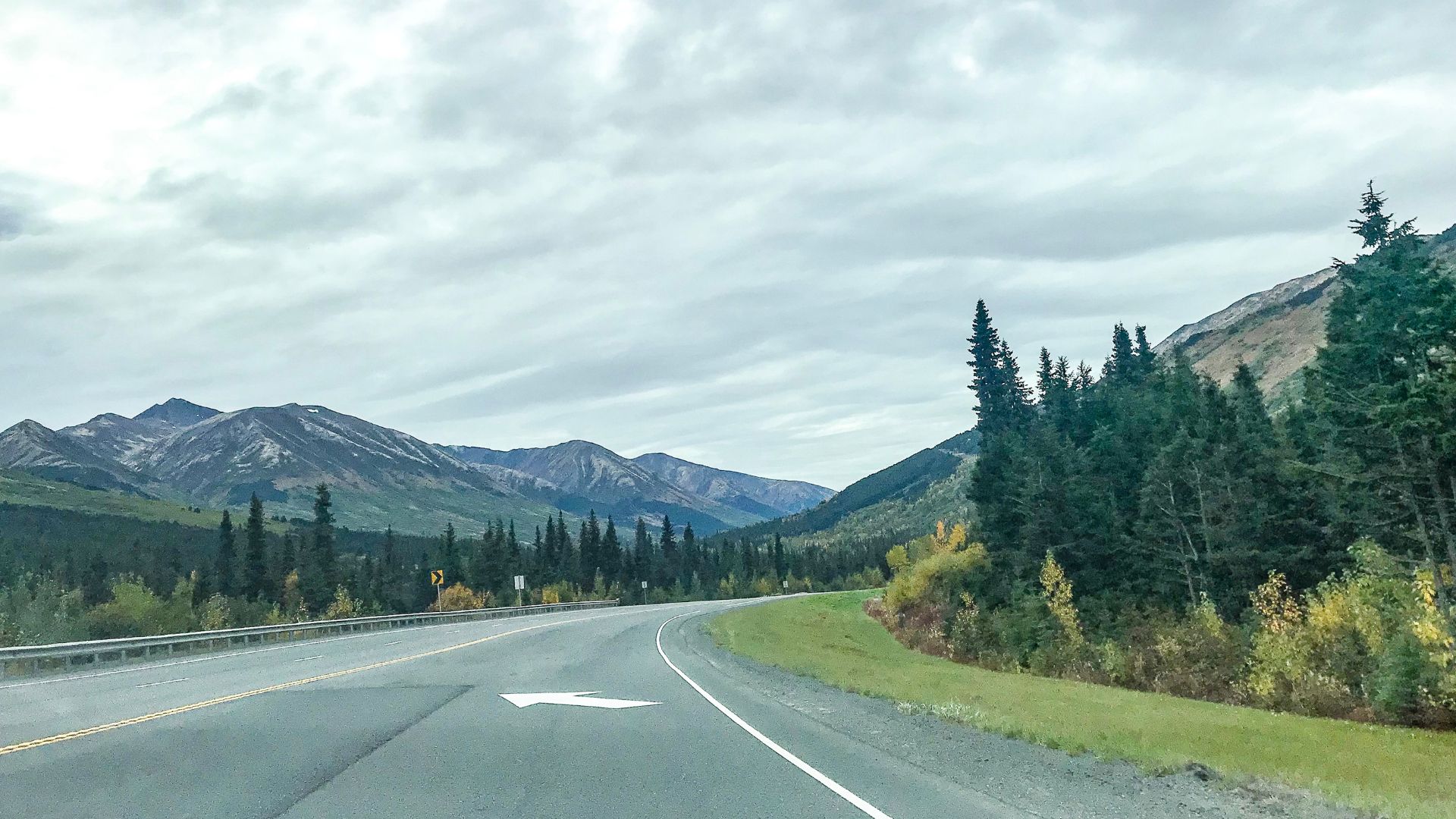 A photo from a moving car on a scenic mountain highway, showing a two-lane road winding through evergreen forests and mountains under a cloudy sky in a northern climate.