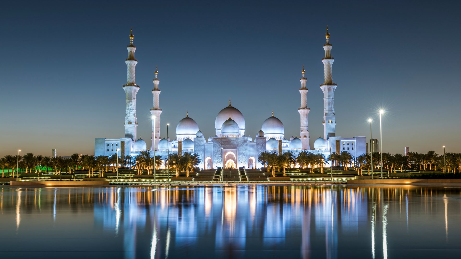 An illuminated white marble mosque with multiple domes and minarets reflects in a body of water at dusk in Abu Dhabi, United Arab Emirates.