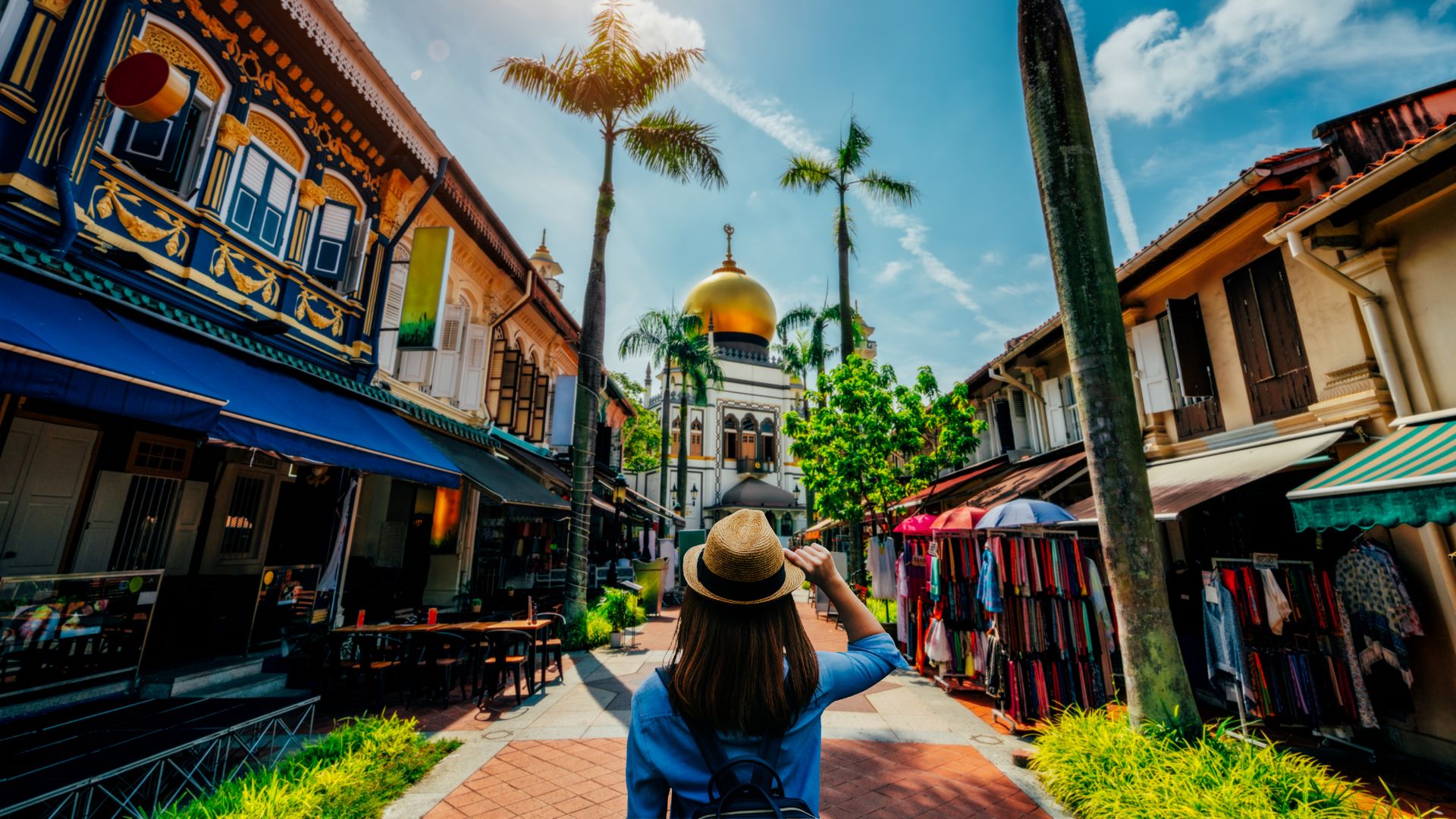 A person in a hat is standing on a pedestrian street lined with colorful shophouses, looking toward the large Sultan Mosque with its prominent golden dome at the far end.
