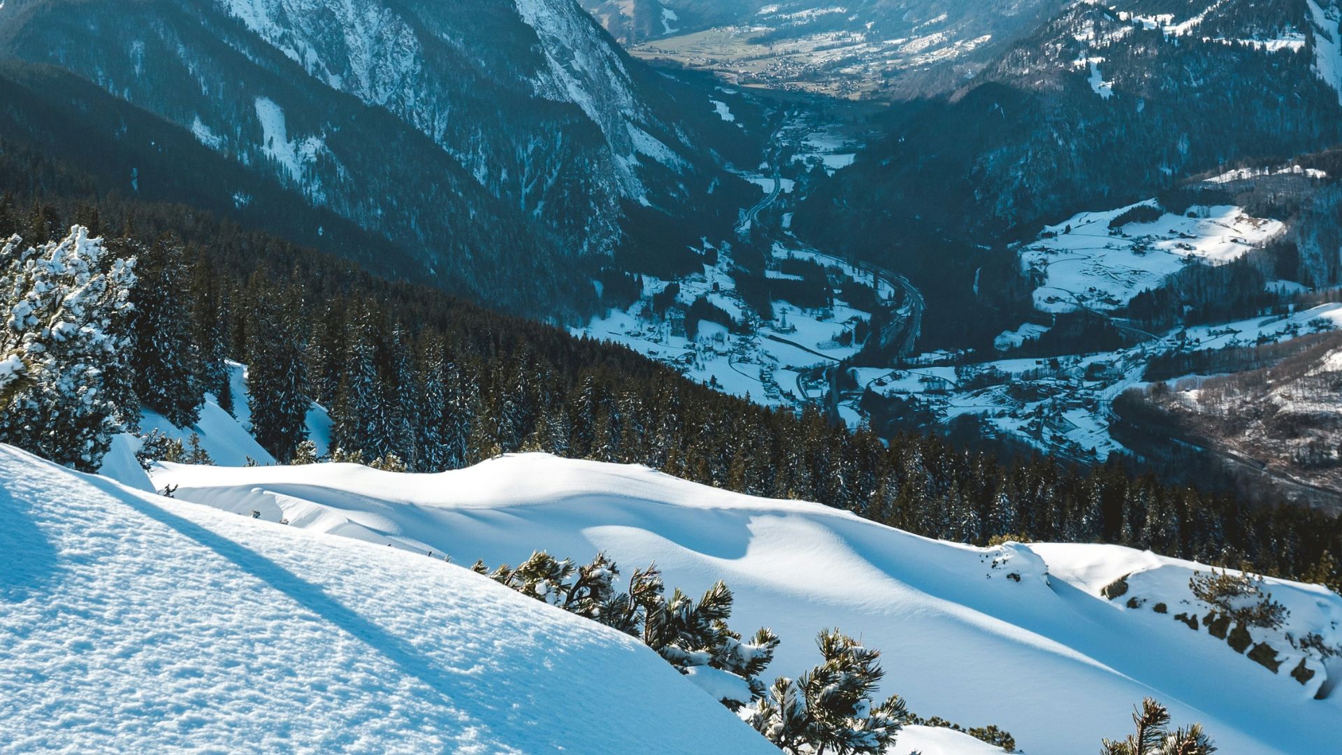 A panoramic view of a vast, snow-covered mountain valley with a small village nestled among pine forests under a bright blue sky.