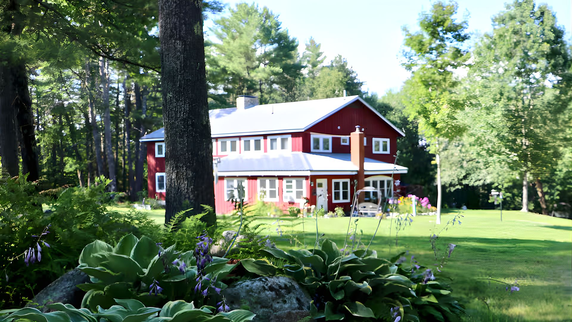 A large, two-story red lodge with white trim, surrounded by a lush green lawn and abundant trees during the daytime in summer.