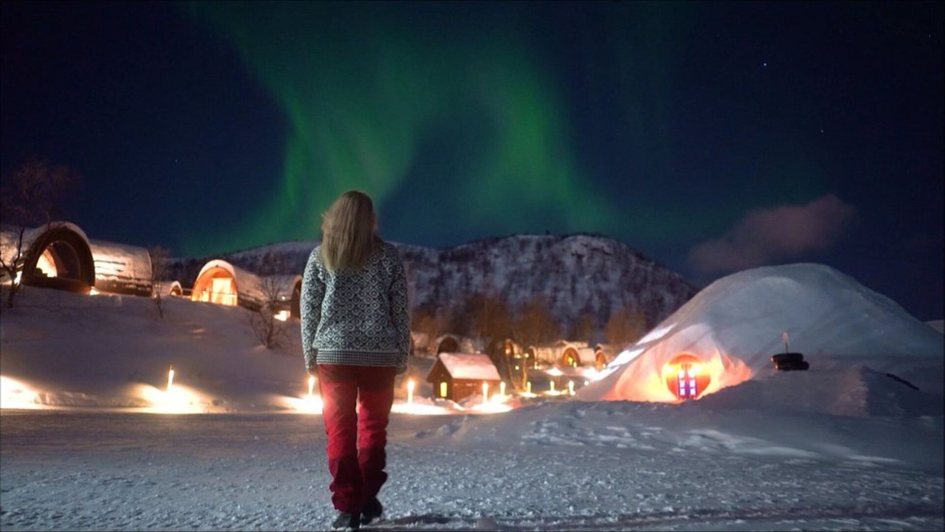 A person in a patterned sweater and red pants stands in the snow, looking up at vivid green Northern Lights in the dark night sky over a snow-covered resort featuring illuminated wooden cabins and a large snow igloo.