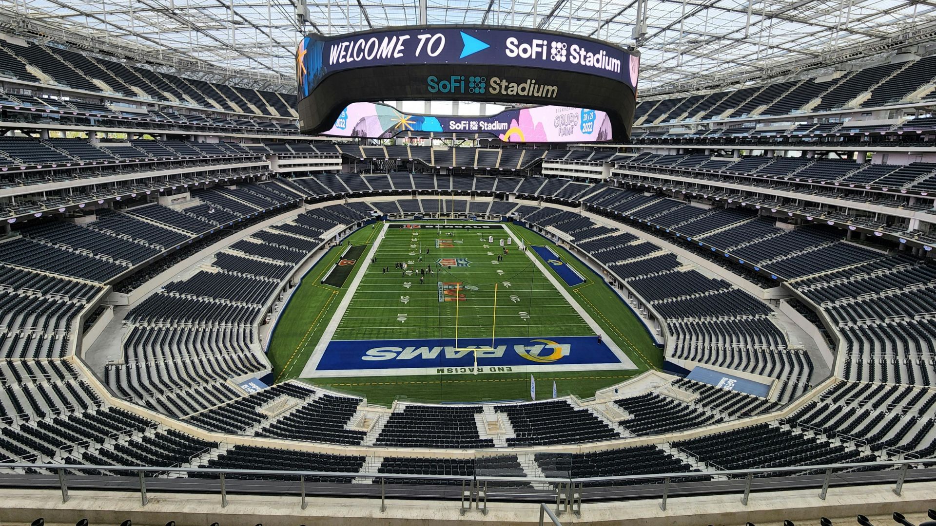 A wide, elevated view of the empty SoFi Stadium interior, showing the massive central video screen, stadium seating, and the Los Angeles Rams logo on the field.