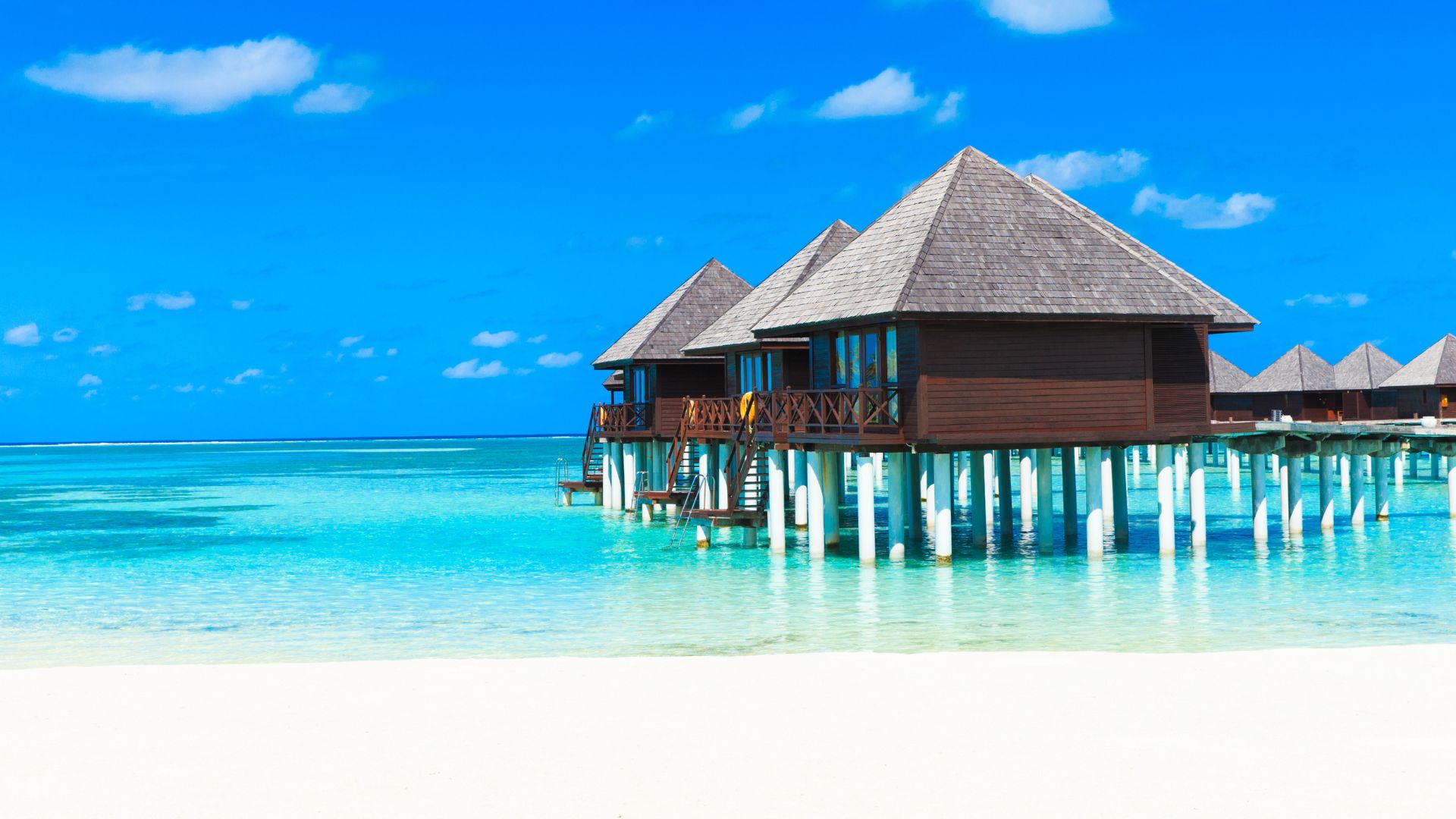 A photograph of several thatched-roof, dark wooden overwater bungalows built on stilts above a shallow, bright turquoise lagoon at the Soneva Fushi resort in the Maldives, with white sand visible in the foreground under a blue sky with white clouds.