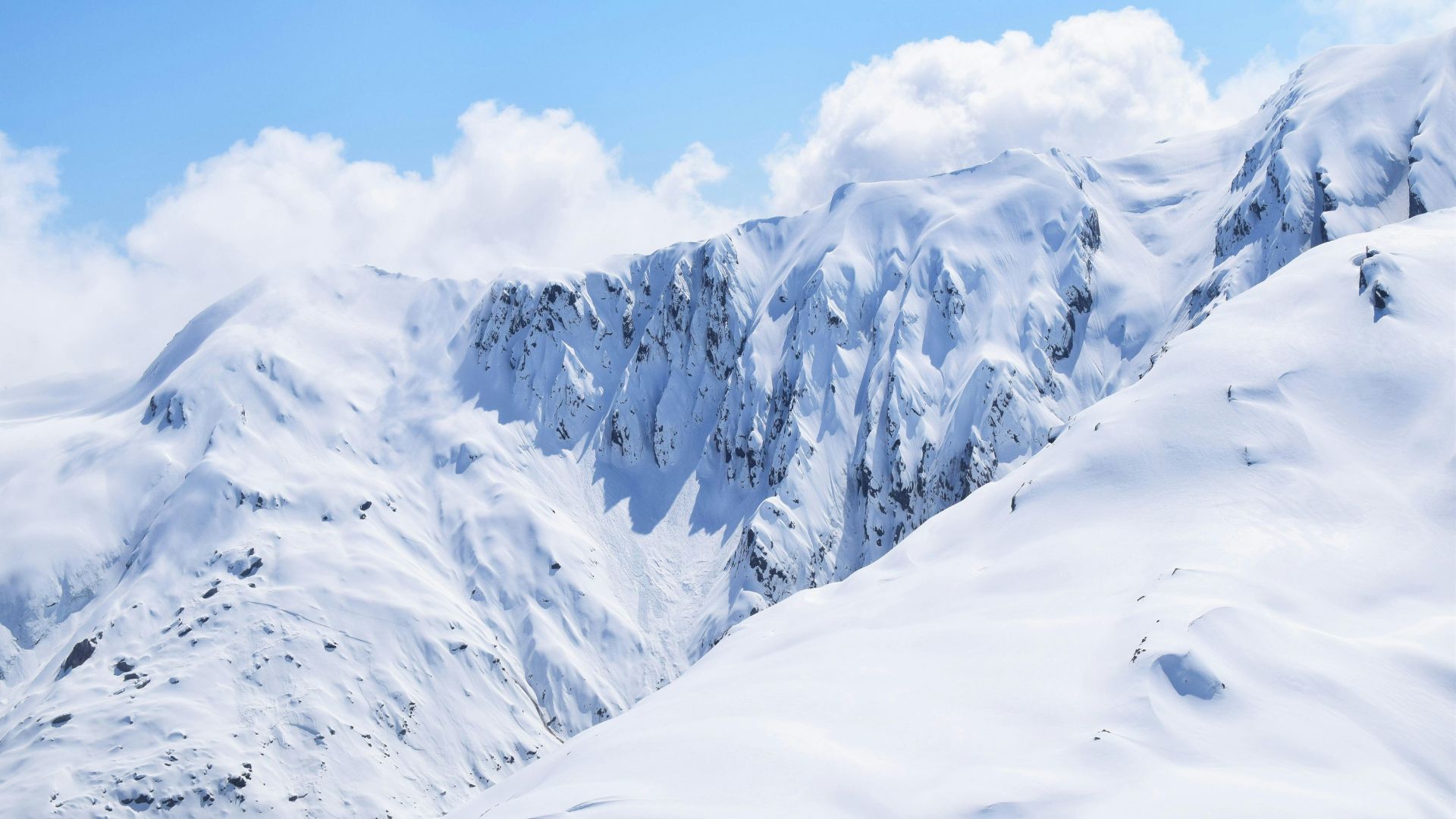 A panoramic view of steep, rugged, snow-covered mountain peaks under a bright blue sky with white clouds.