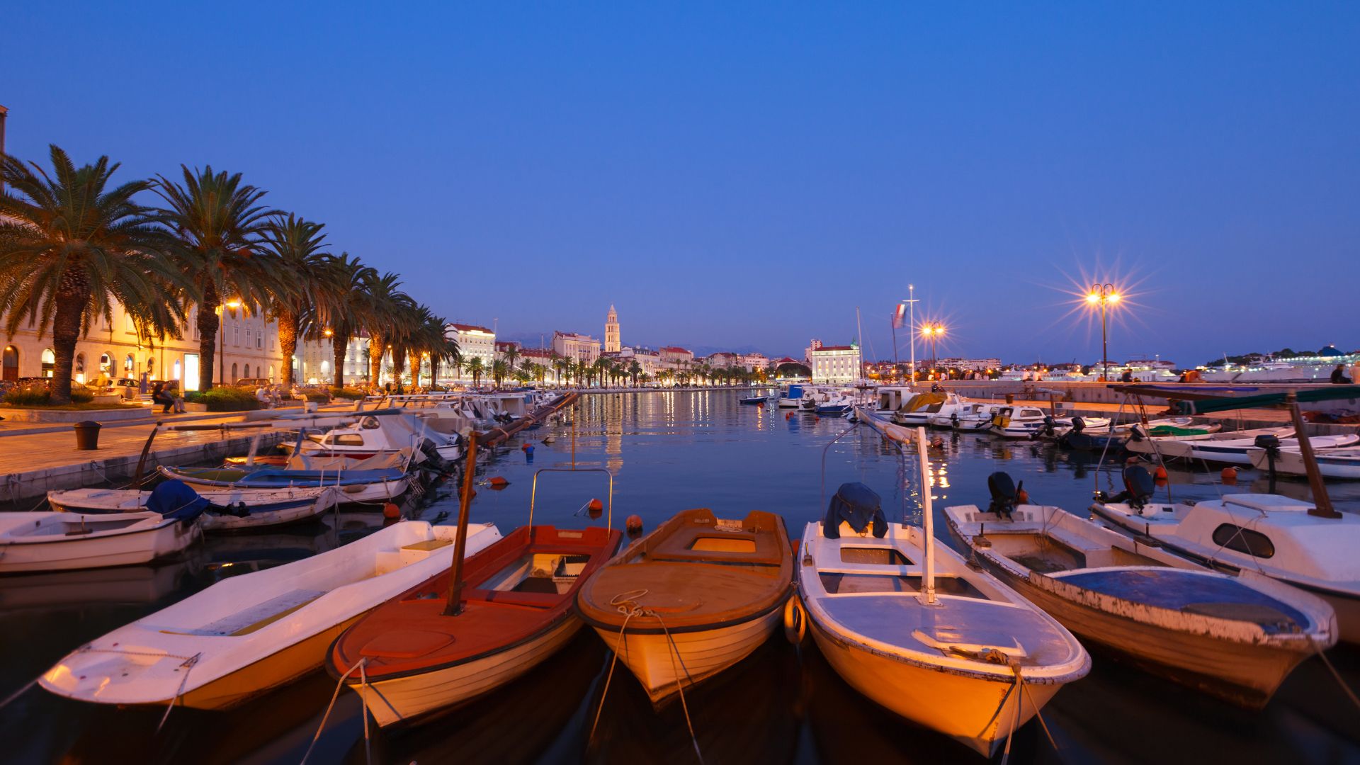A twilight photograph of a calm harbor with several small boats moored in the foreground, lined by a palm-tree-fringed promenade and a lit-up historic cityscape with a bell tower in the distance.