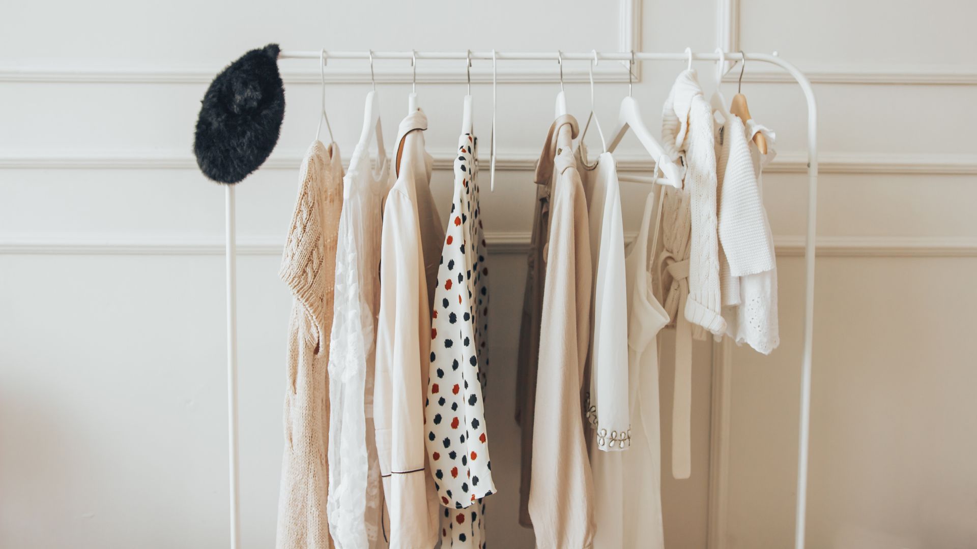A minimalist capsule wardrobe on a white metal rack, featuring various tops and shirts in neutral colors and a single black hat.