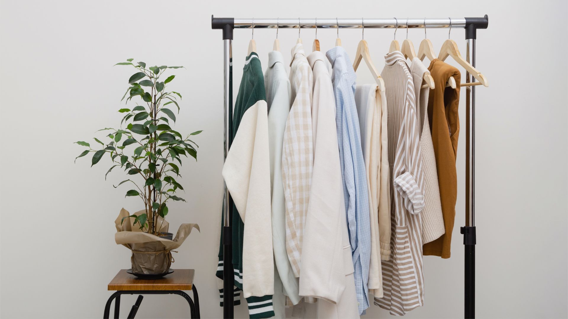 A clothing rack filled with an assortment of neutral and green tops, shirts, and sweaters, with a potted green plant on a small wooden stool beside it.