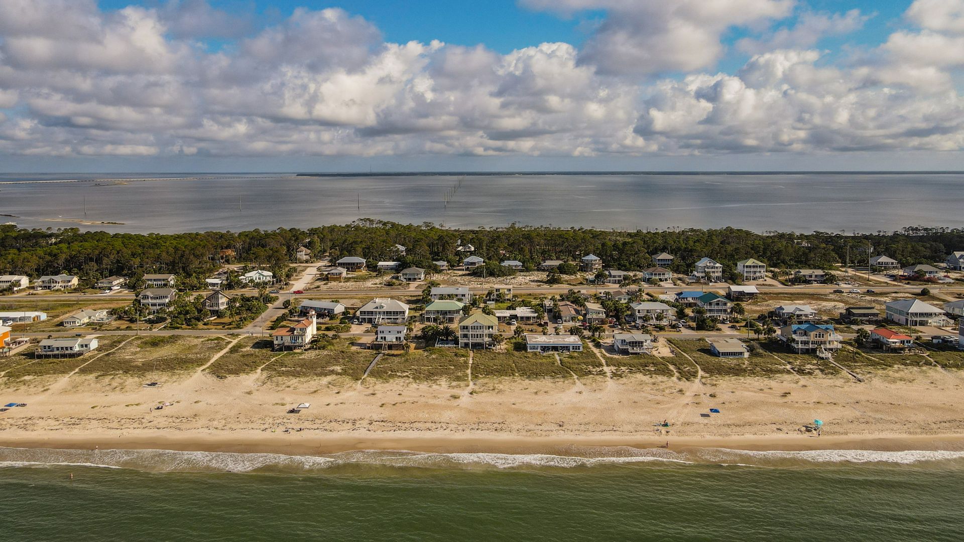 An aerial photograph of a sandy barrier island lined with beach houses and green dune grasses, with ocean waves on one side and a calmer bay visible in the distance under a partly cloudy sky.
