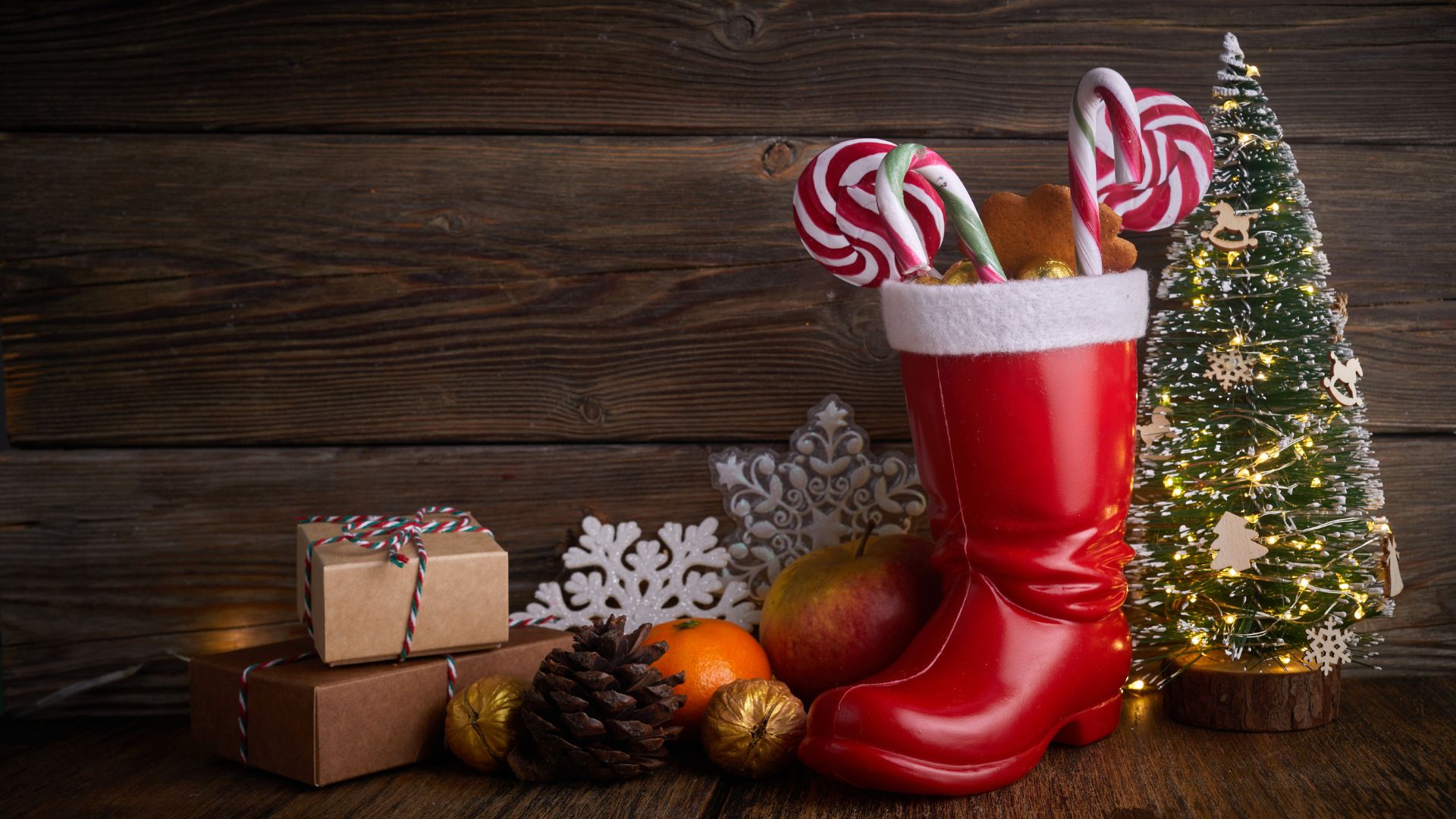 A red Santa boot filled with candy canes and small treats, surrounded by small gift boxes, an orange, an apple, pinecones, and a small, lit Christmas tree against a rustic wooden background.