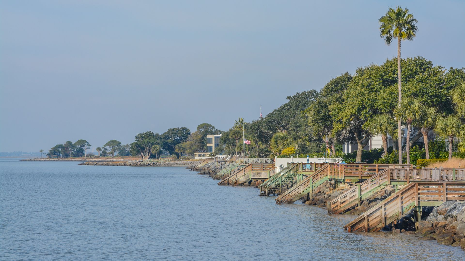 A coastal scene in St. Simons Island, Georgia, featuring a concrete and rock seawall with several wooden stairways descending to calm blue water, with palm trees and dense green foliage on the high bank in the background.